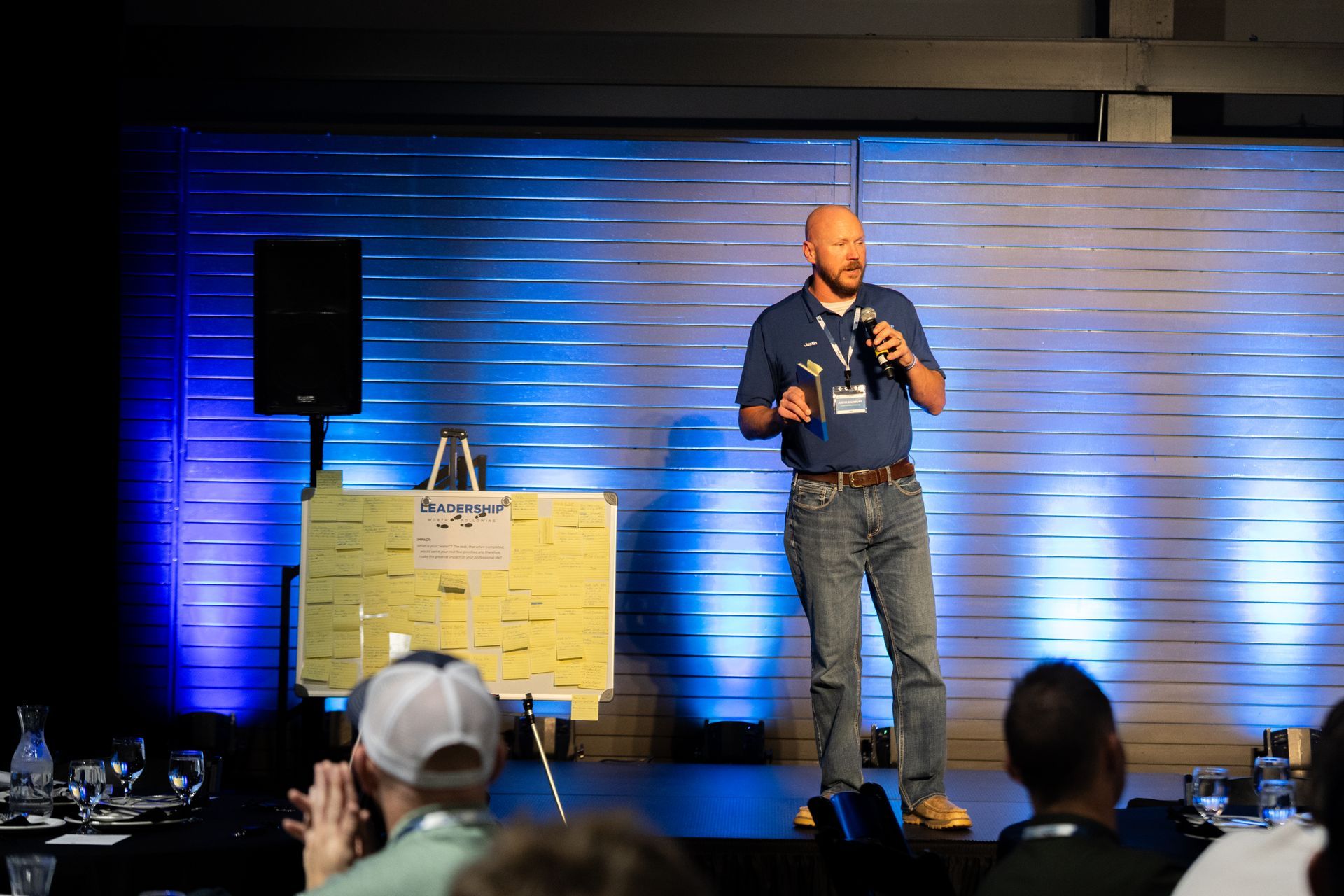 Man speaking on stage, holding a microphone. Audience seated, applauding at Leadership Worth Following car wash convention