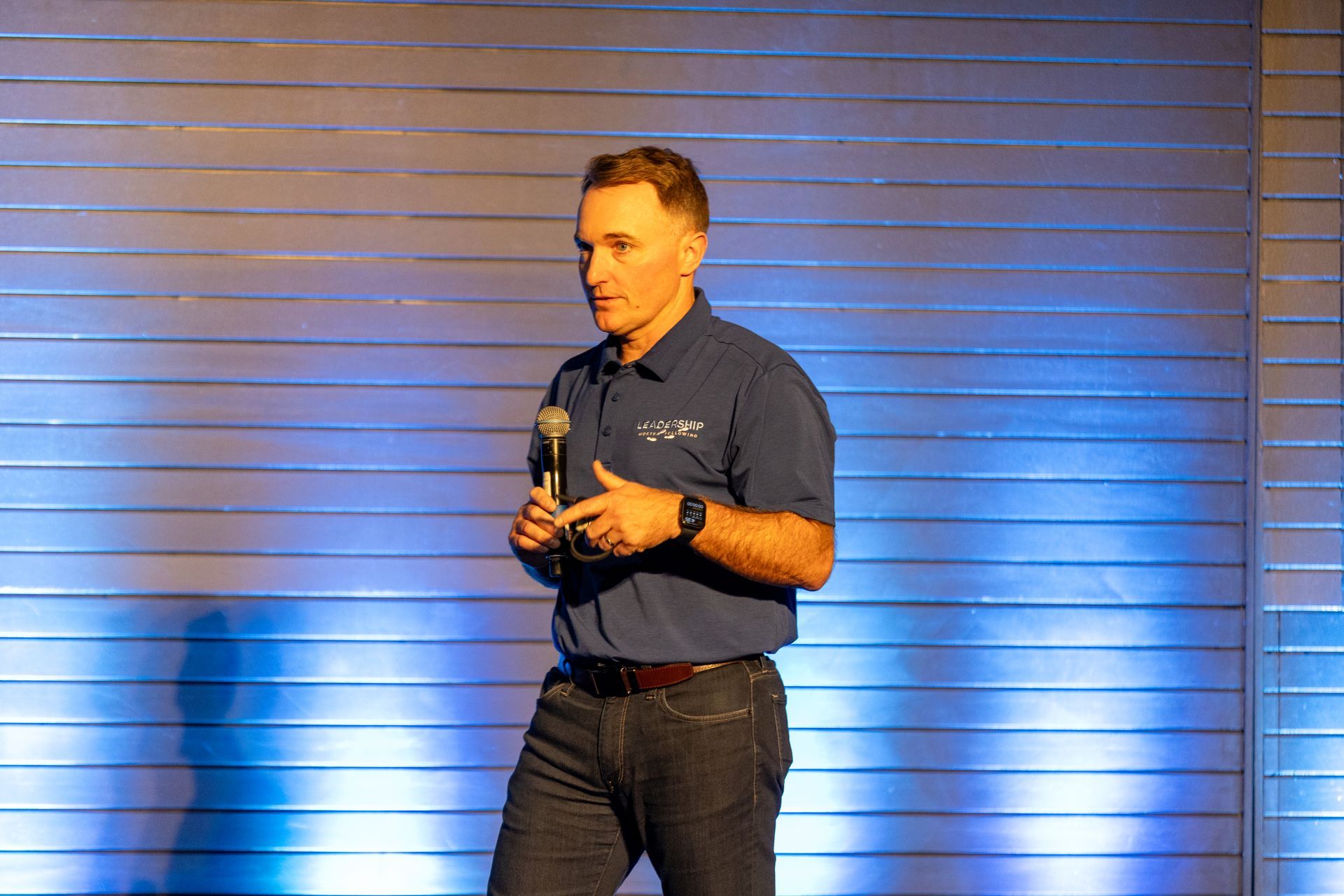 Matt DeWolf speaking at Leadership Worth Following car wash convention, holding a microphone on a stage with a blue background.