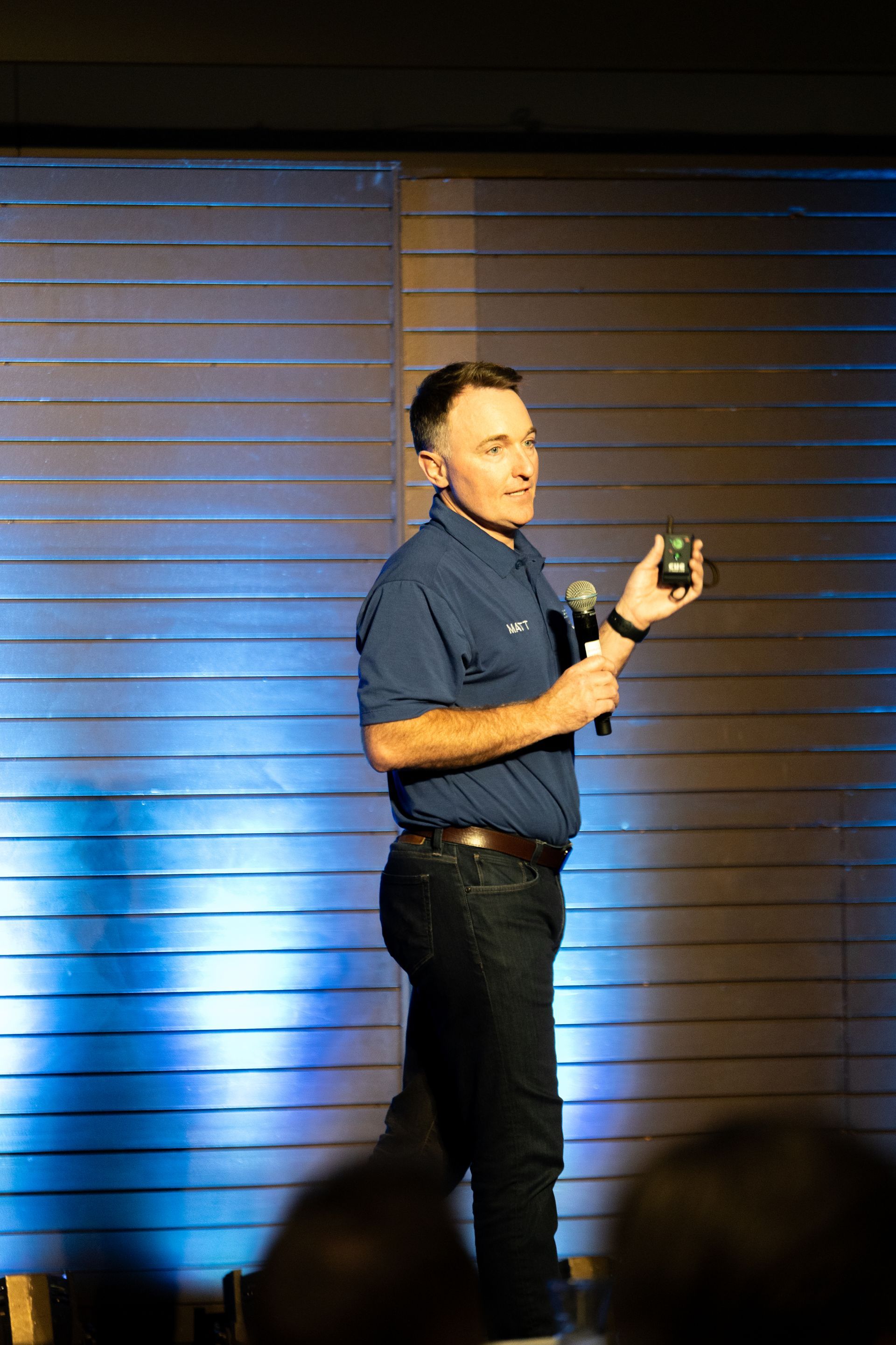 Man on stage at Leadership Worth Following car wash convention. He wears blue shirt, jeans. Background: wood wall, blue light.