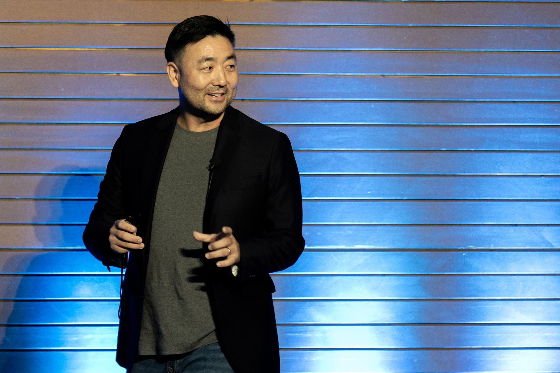 Man in blazer, grey shirt, gesturing onstage, smiling, speaking into a microphone at Leadership Worth Following car wash convention