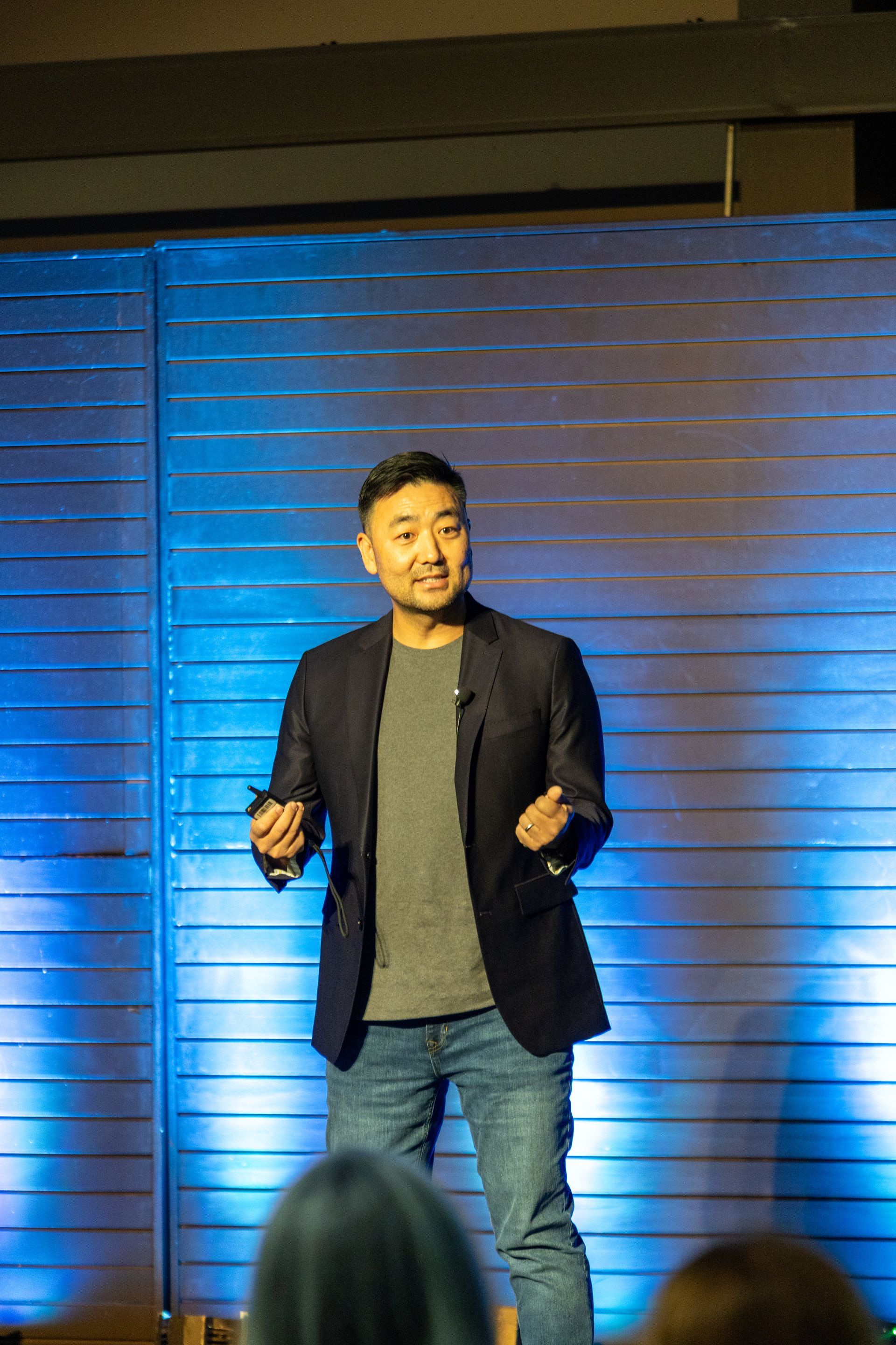 Man in a blazer and jeans gestures while speaking on a stage with a blue-lit backdrop.