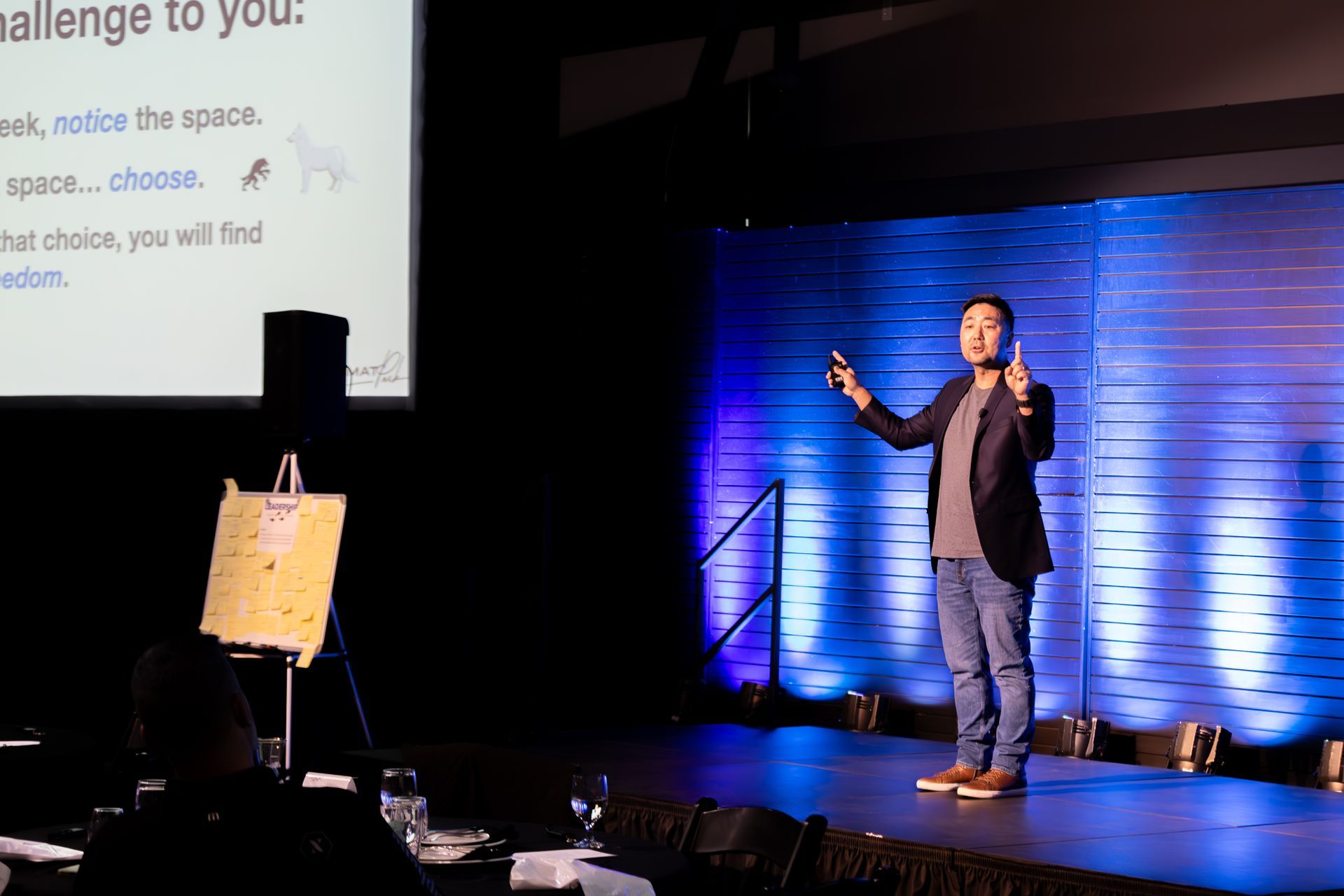 Man giving a presentation at Leadership Worth Following car wash convention. Dark suit jacket, jeans. Blue and gold stage lighting.