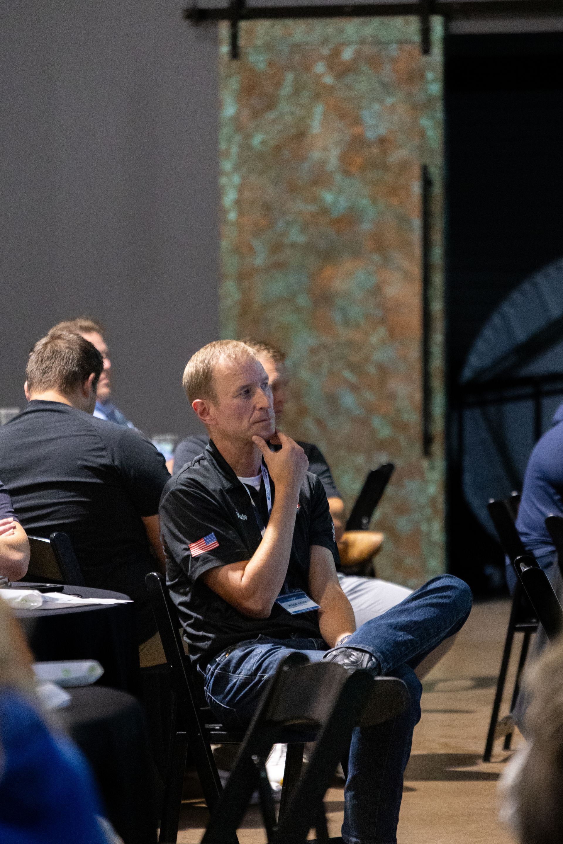 Man in black shirt and jeans sits thoughtfully, hand on chin, at an event.