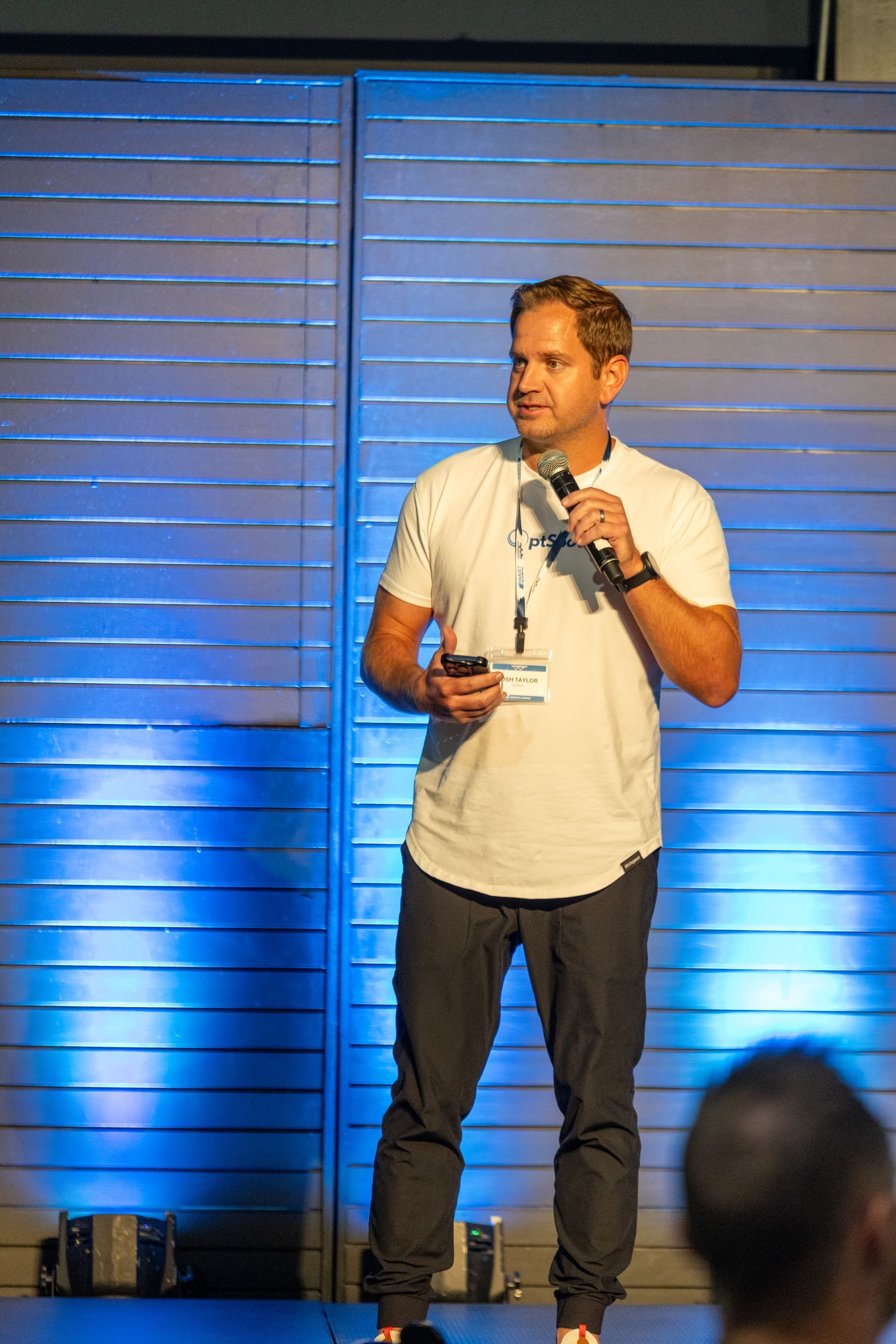 Man speaking at a conference, holding a microphone and small device. Blue lit wall behind him.