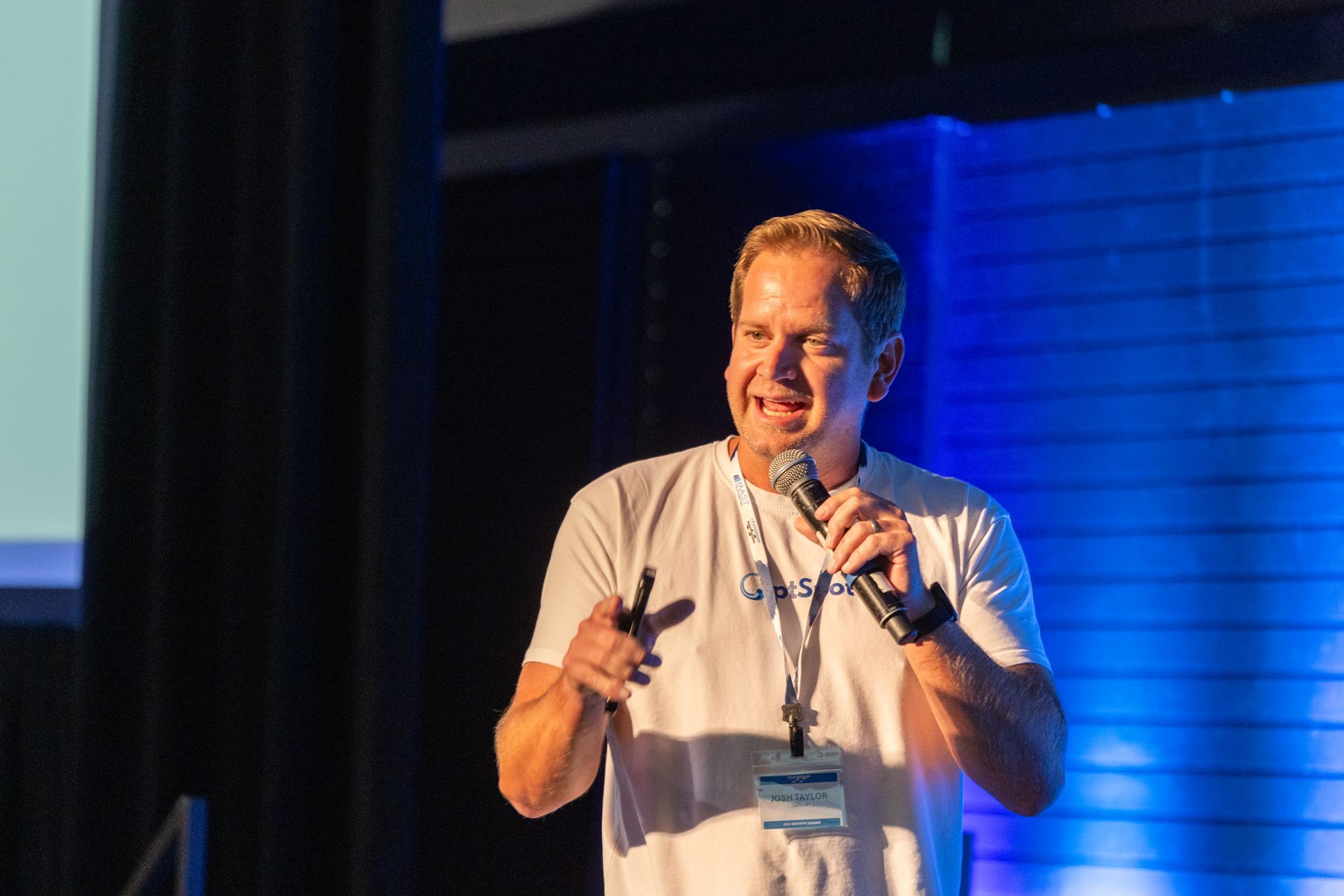 Man in white t-shirt speaking into a microphone on stage, gesturing with his hands. Blue and white backdrop.