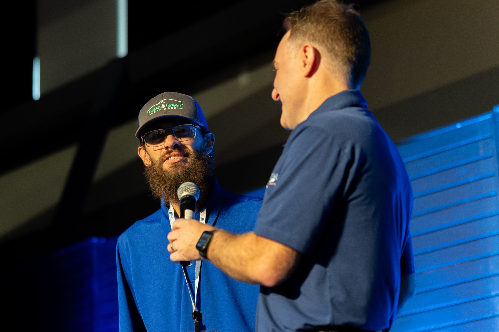 Two men on stage, one with a beard holding a microphone, the other smiling.