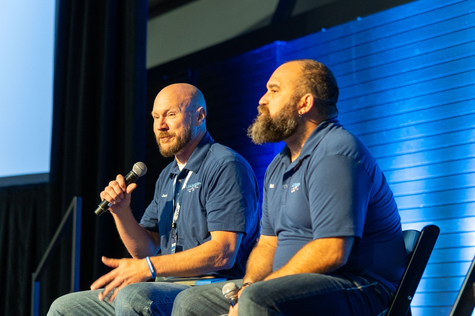 Two men in blue shirts on stage, one speaking into a microphone. Blue background.