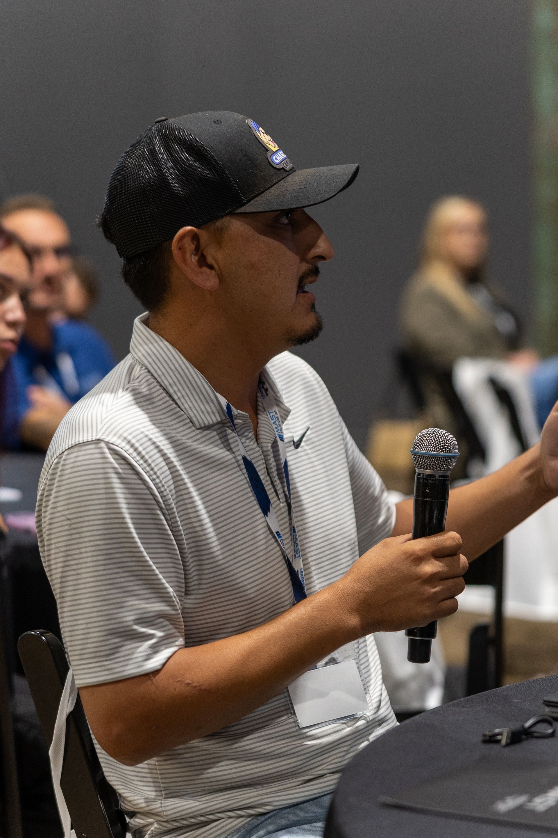 Man in cap and polo shirt speaking into a microphone at an event, gesturing with his hand.