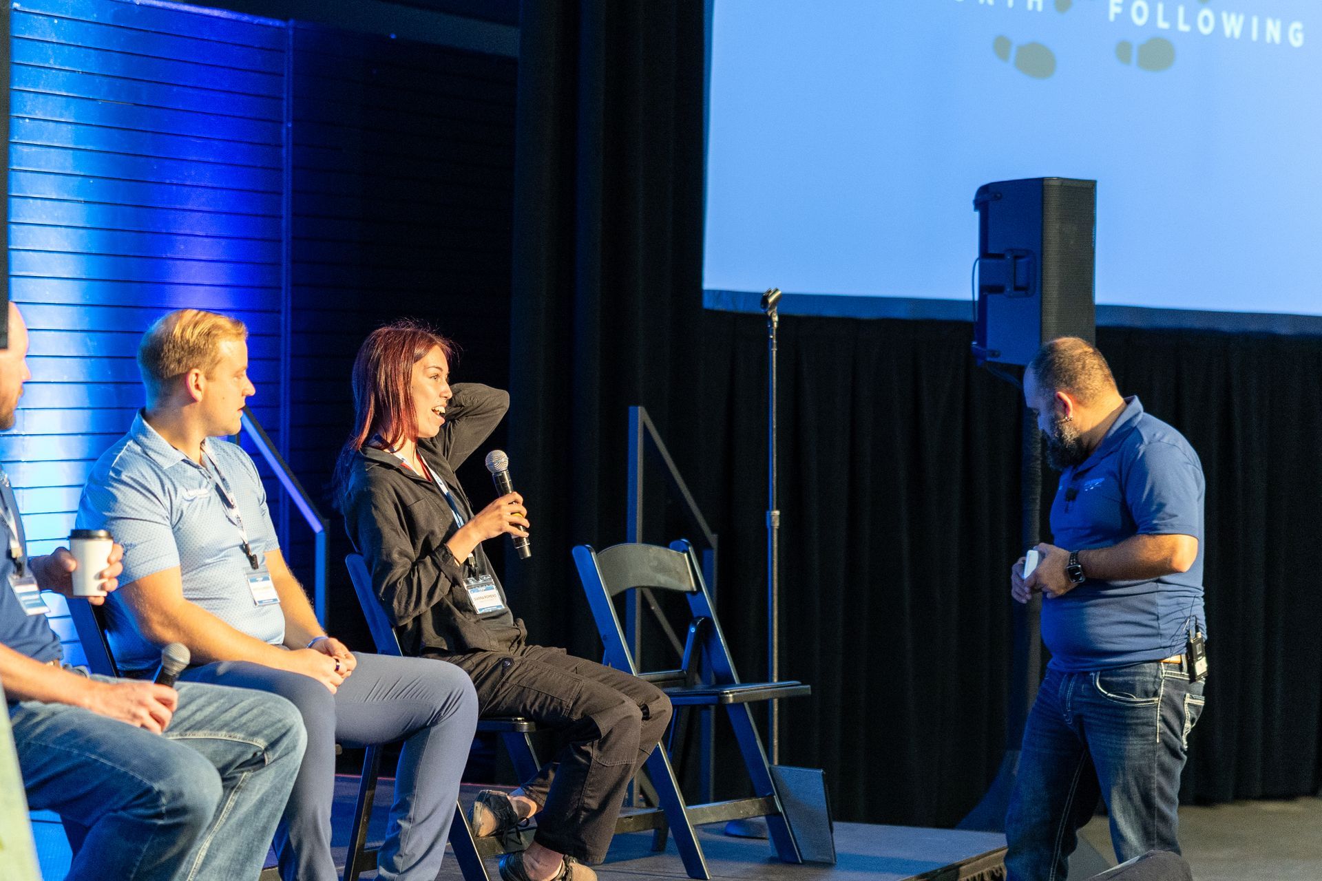 Panel discussion onstage; four people seated, one speaking. Stage with blue backdrop and screen. Man in blue shirt standing nearby.