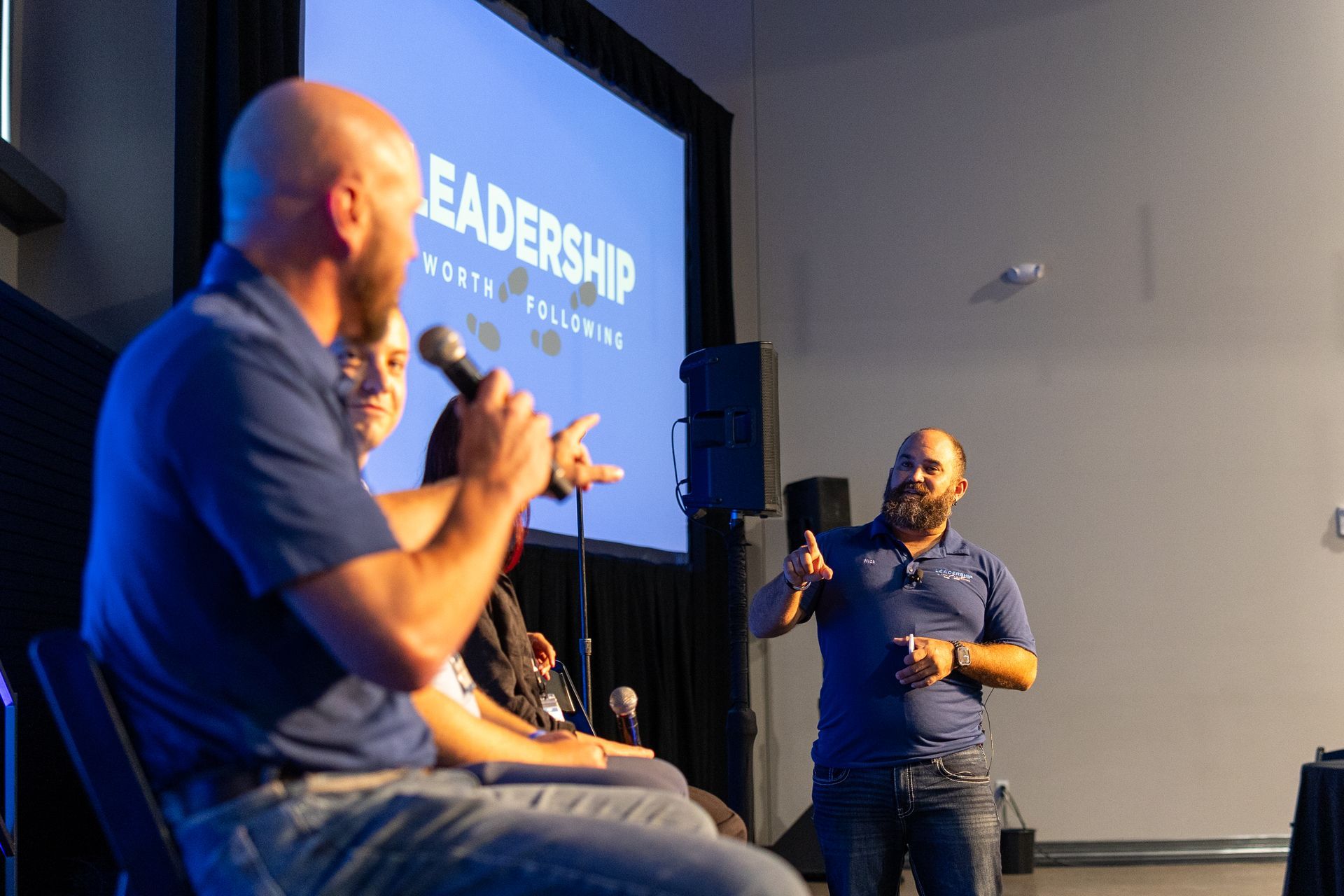 Panel discussion: Three people on stage, one holding a mic. A man stands, gesturing.