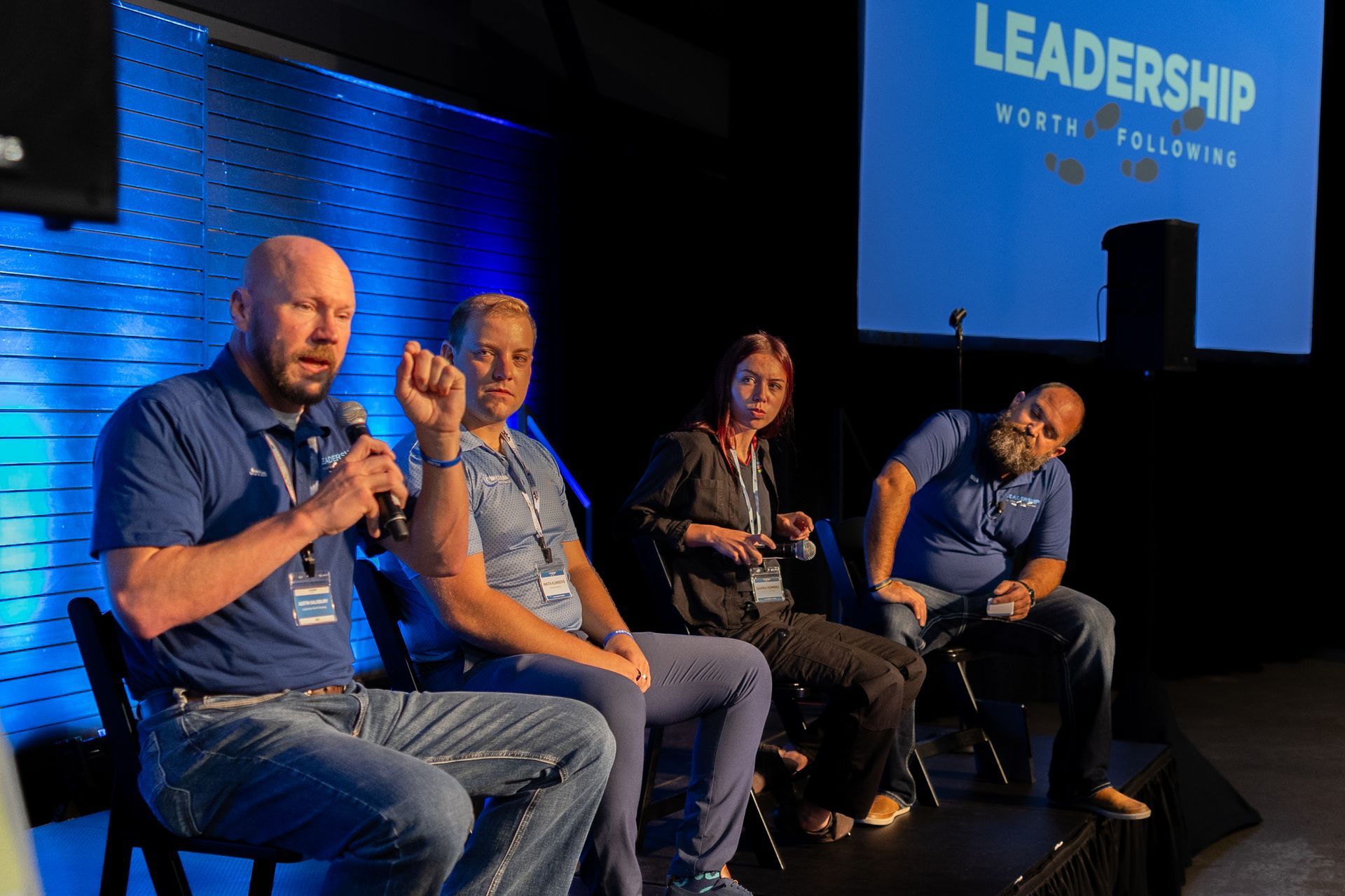 Panel of four people seated on a stage, discussing leadership. One speaks into a microphone. Blue background and text.