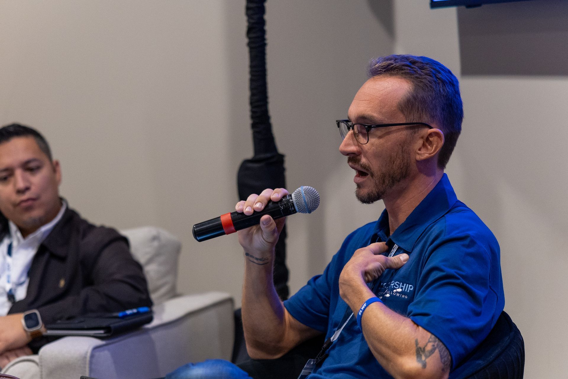 Man with glasses speaking into a microphone; another man sits next to him.