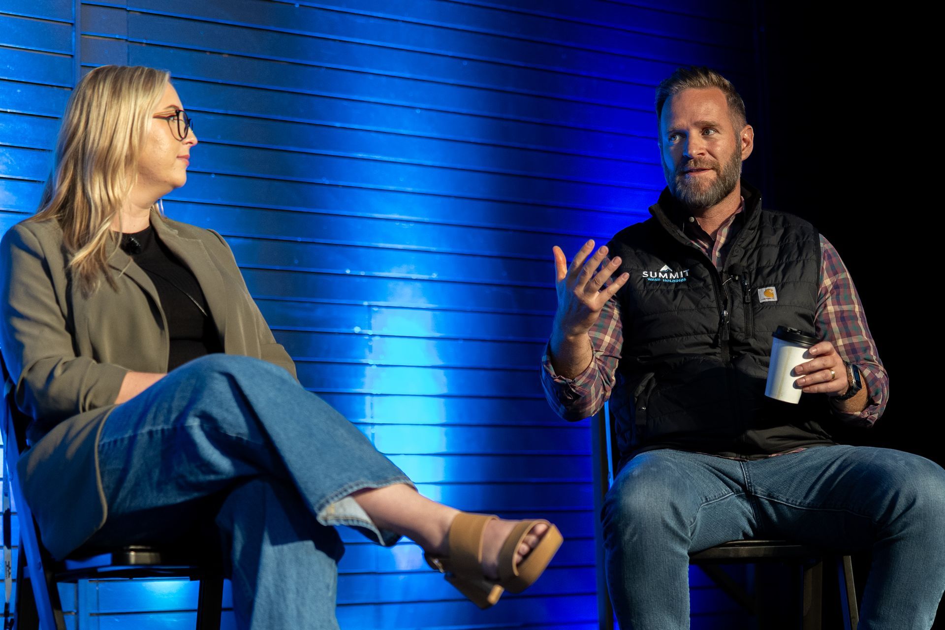 Two people on stage: a woman in glasses and jeans, a man in a vest. Both seated, conversing, with blue backdrop.
