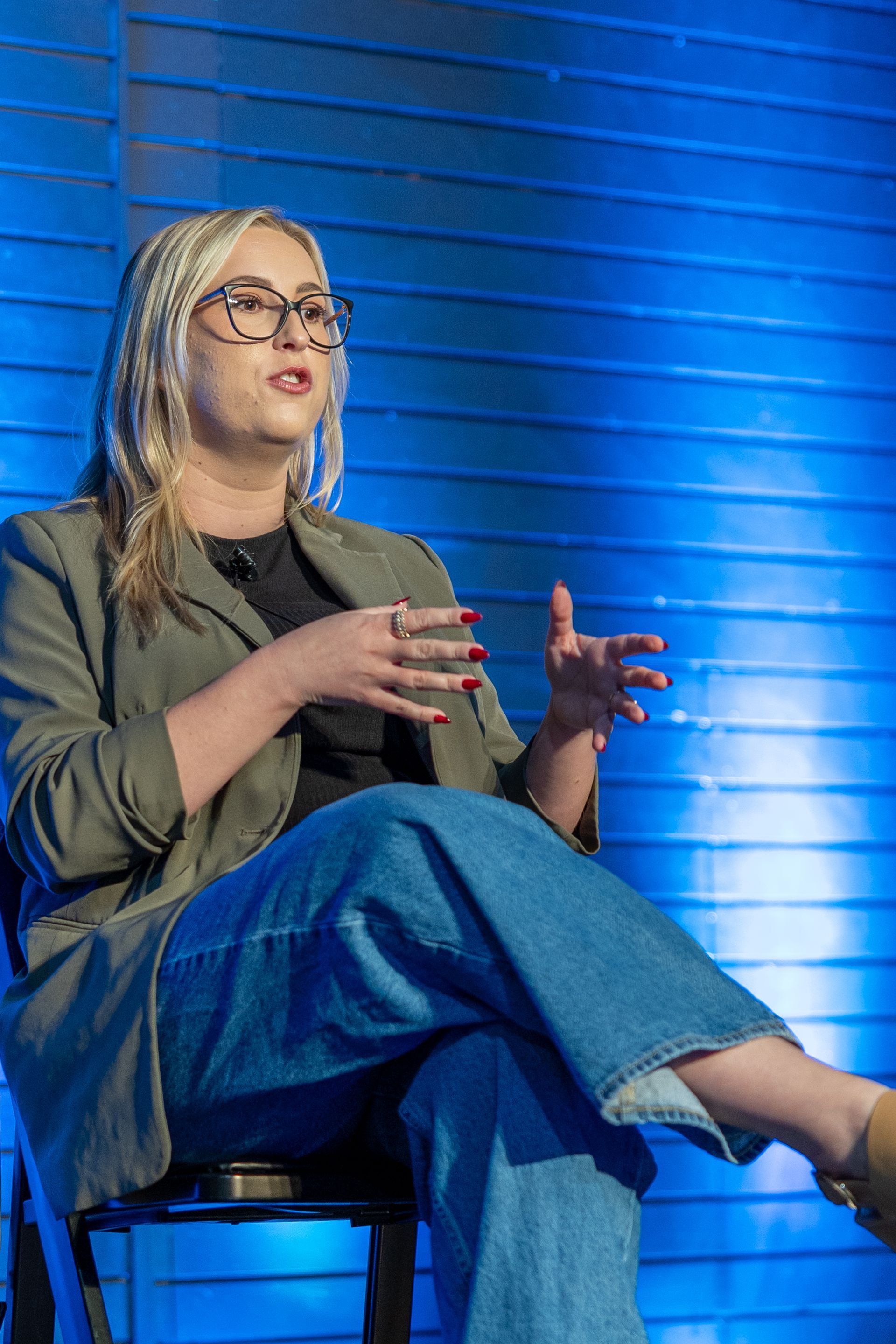 Woman with glasses, gesturing while seated. Wearing denim jeans and blazer against a blue panel backdrop.