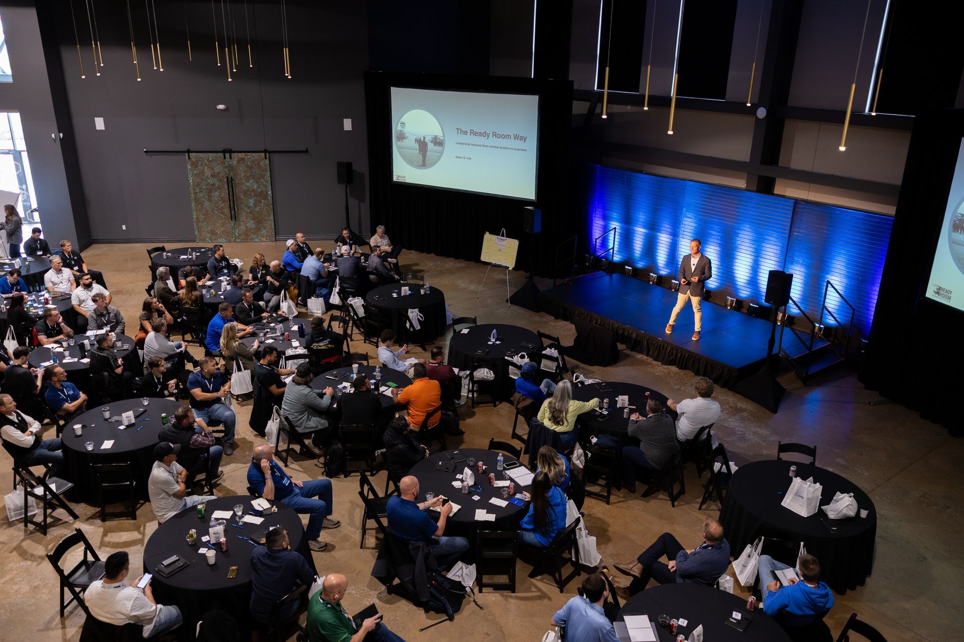 Audience at tables watches a speaker on stage at an event in a large hall.