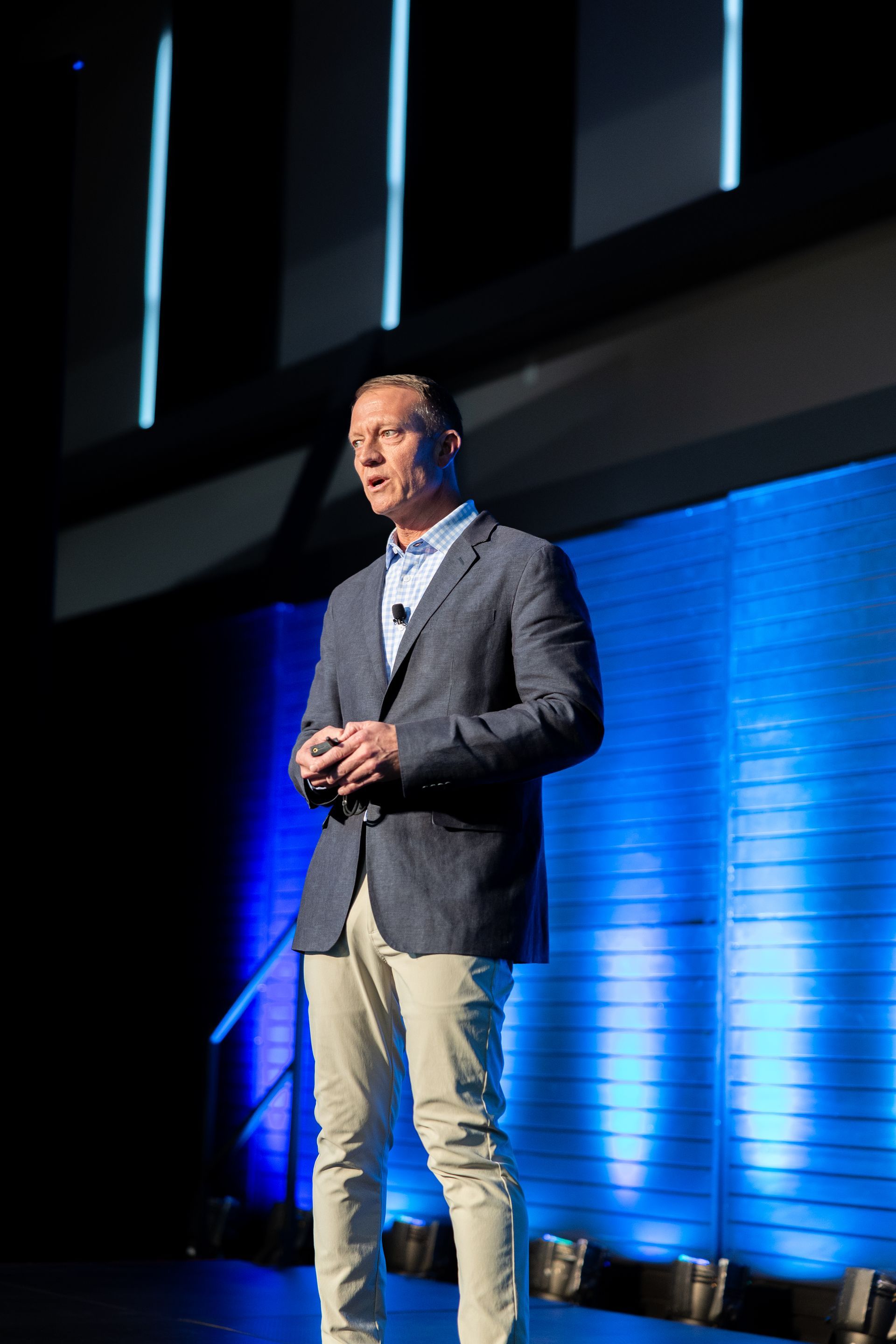 Man in a blazer and tan pants on stage, speaking. Blue and white stage lighting.