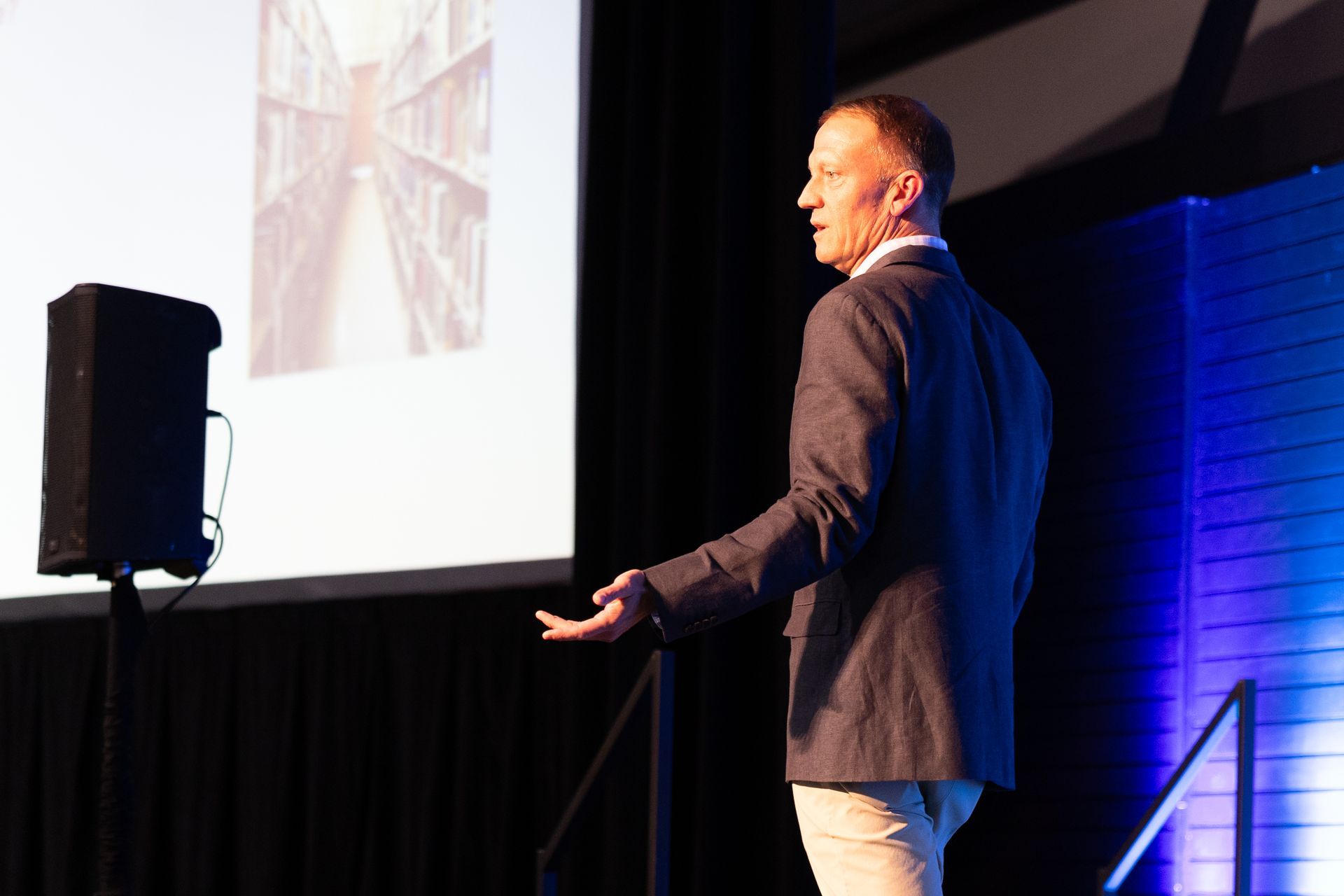 Man giving a presentation, gesturing towards a projected image of a library, in a lit auditorium.