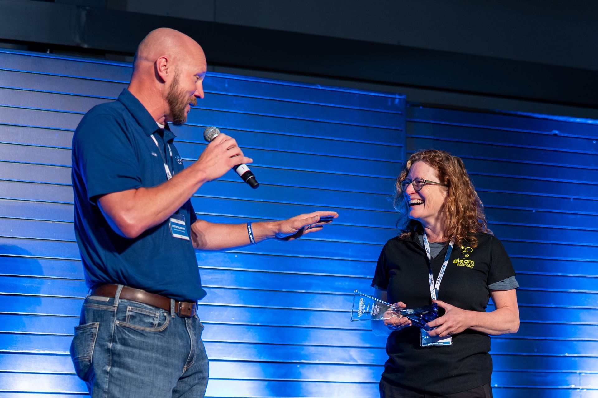 Man with microphone gestures toward laughing woman on stage. Blue and black clothing.