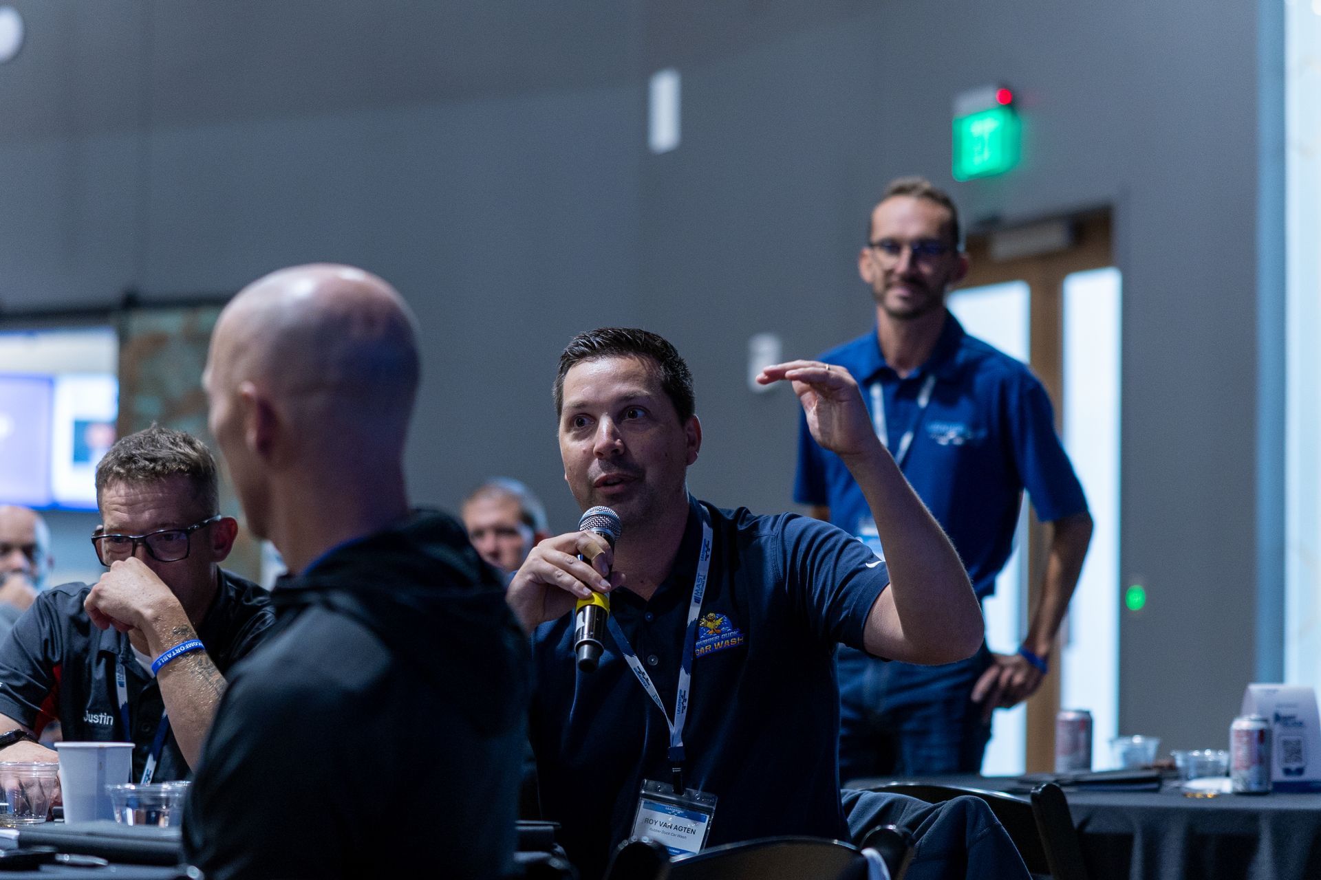 Man speaking into microphone, gesturing during a conference, other attendees listening.