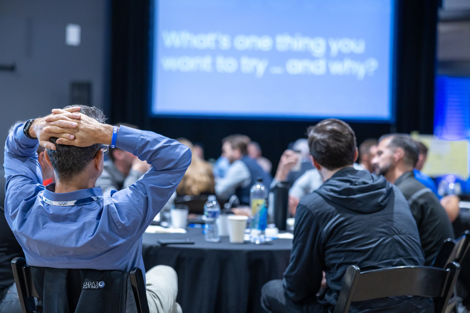People at a conference, seated around tables, facing a screen with text,