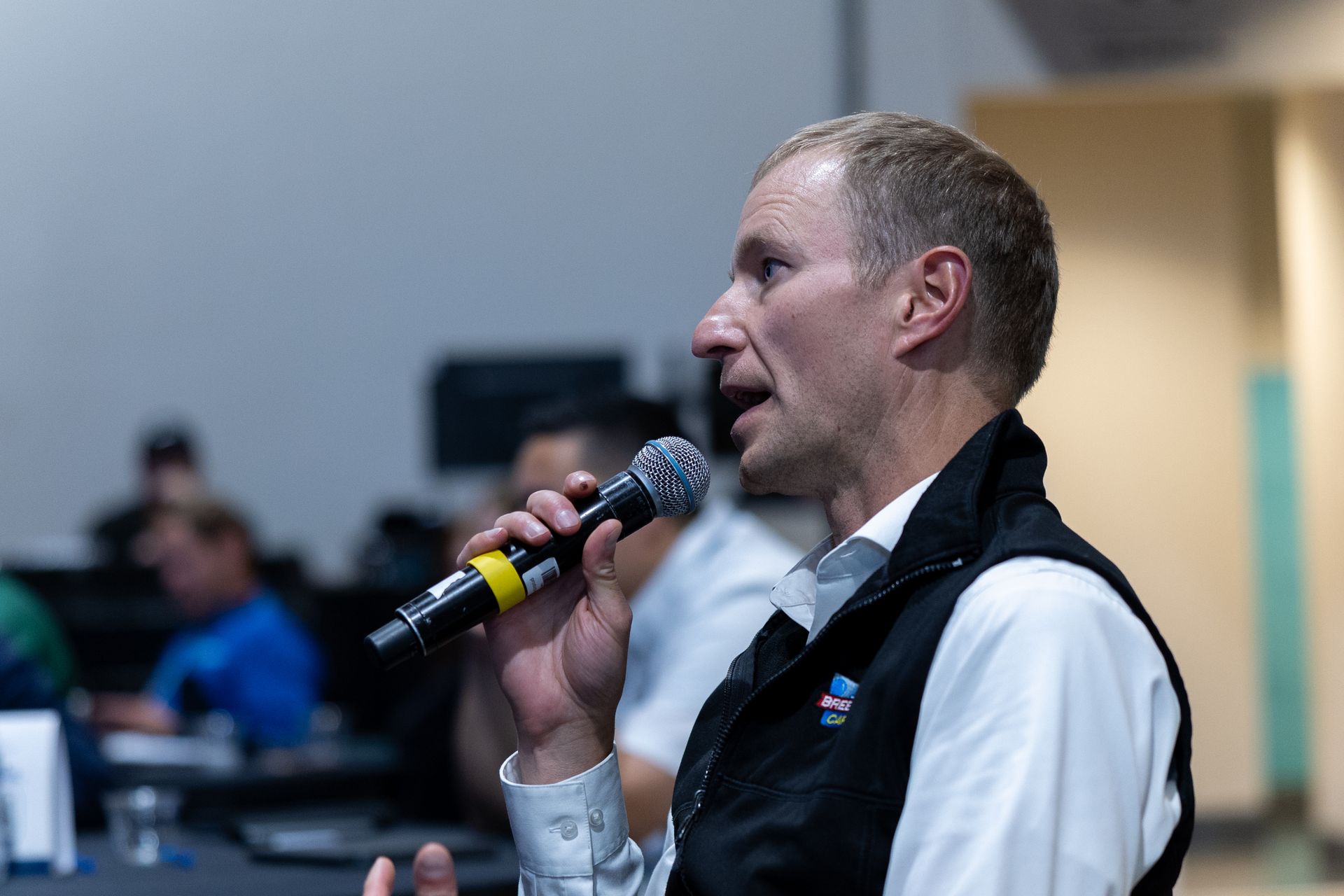 Man speaking into a microphone, wearing a vest and collared shirt, in a room with other people.