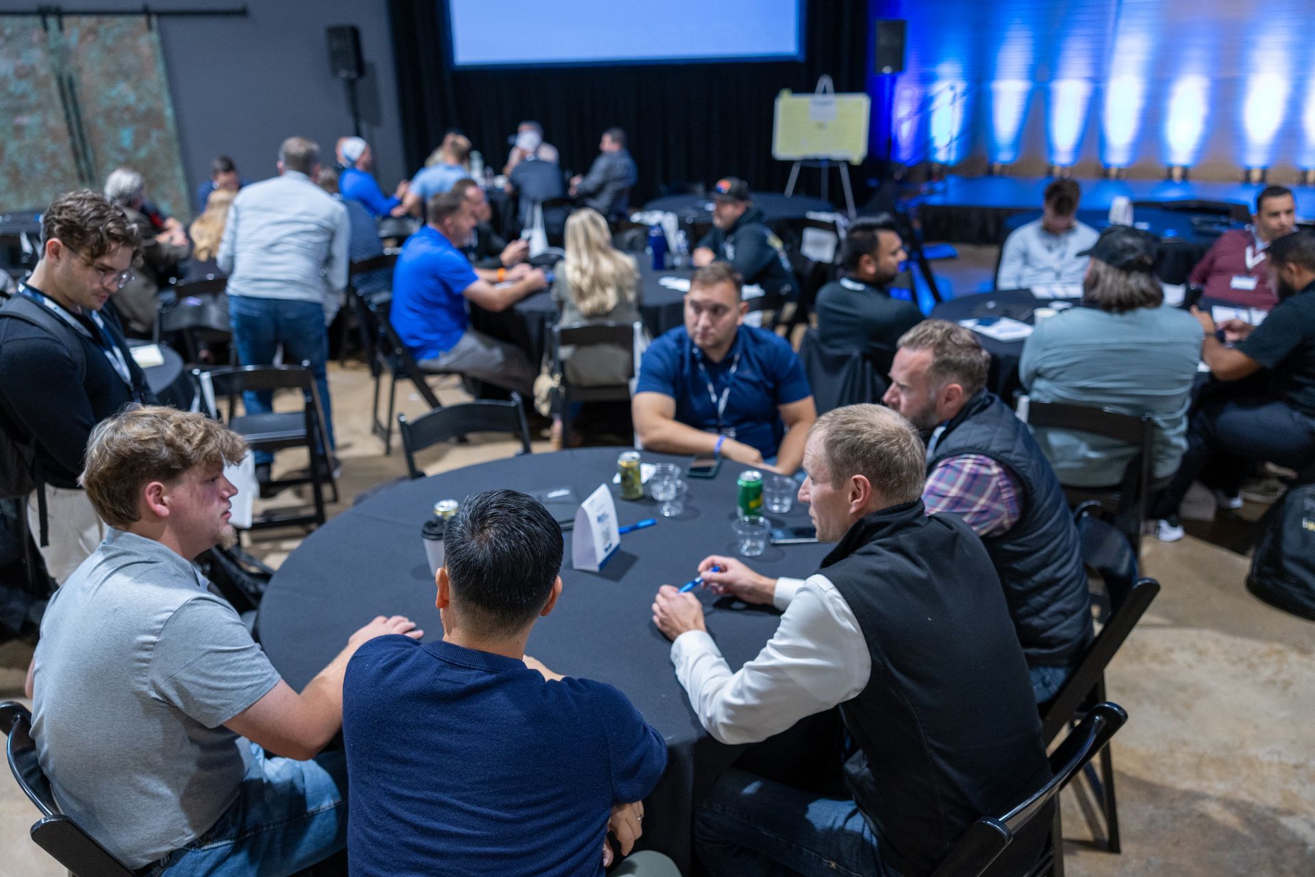 People seated at round tables in a conference hall, engaged in conversation. Dim lighting, dark chairs, and a screen in the background.