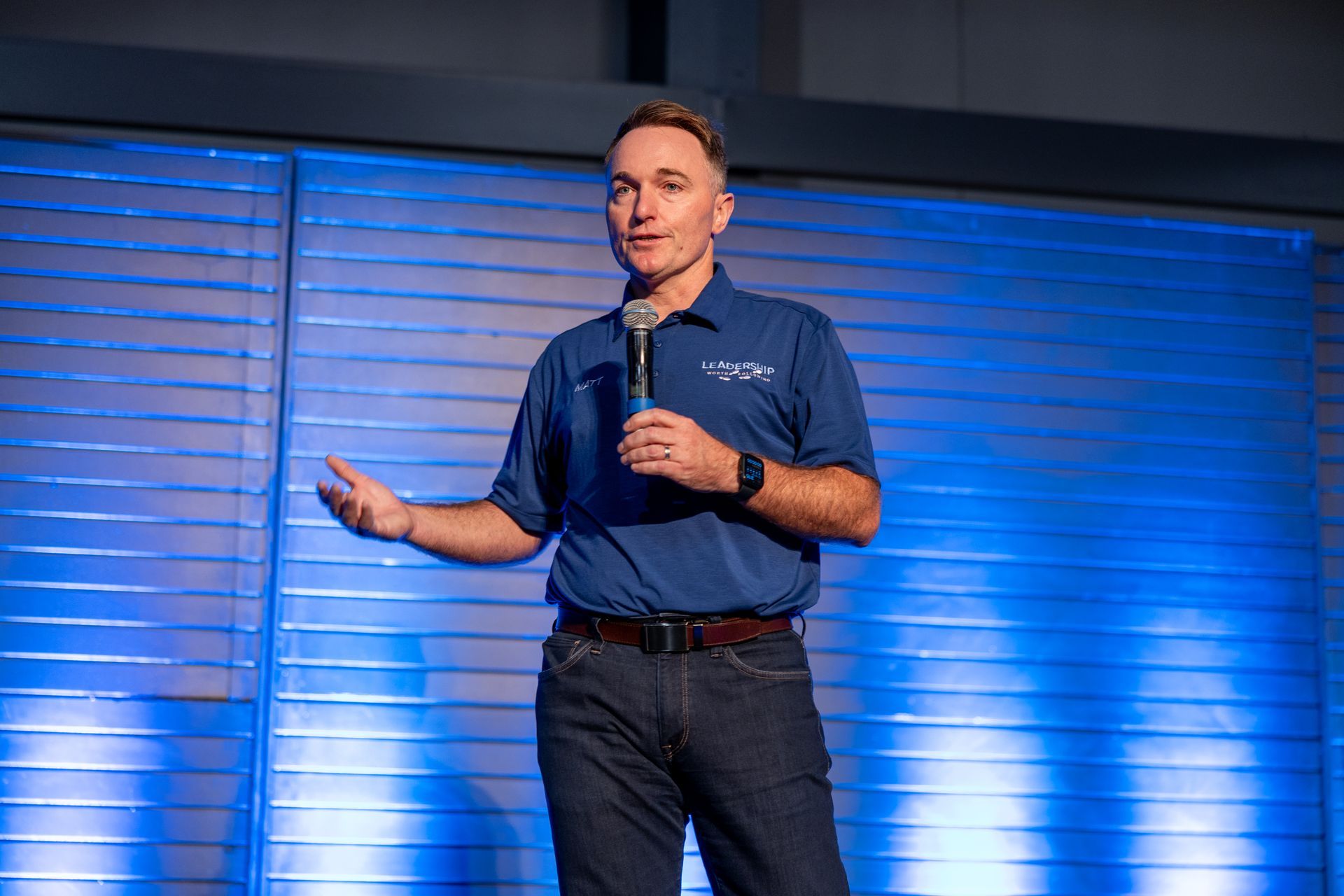 Man in blue shirt speaks into microphone at a conference; blue background.
