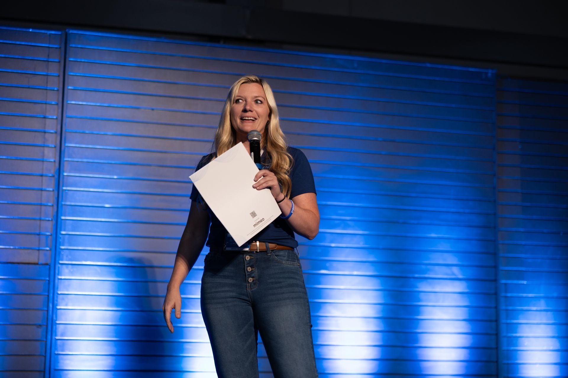 Woman on stage with microphone, holding paper, speaking. Blue background with stage lighting.