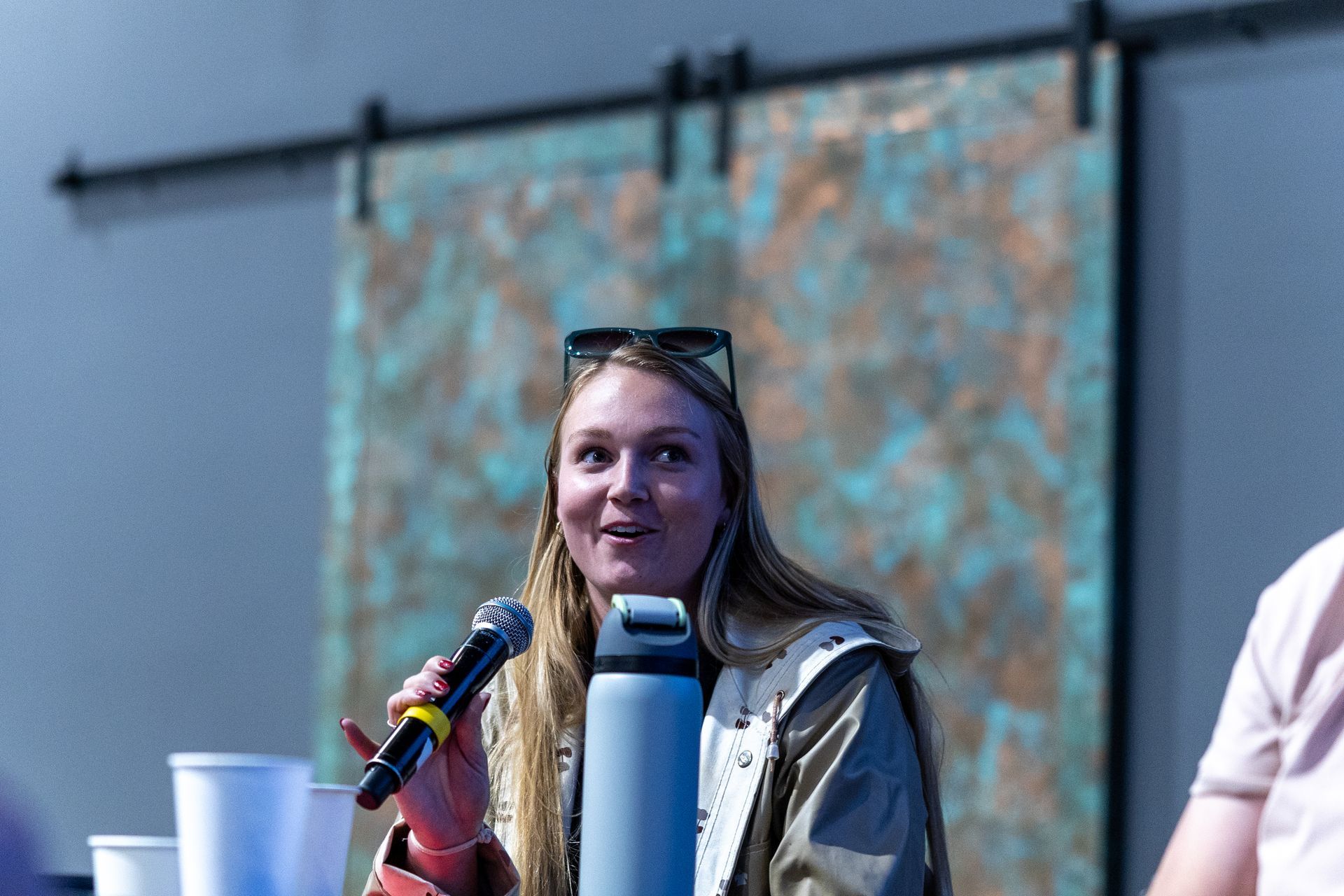 Woman speaking into a microphone, wearing sunglasses and a jacket. Light blue bottle, light setting.