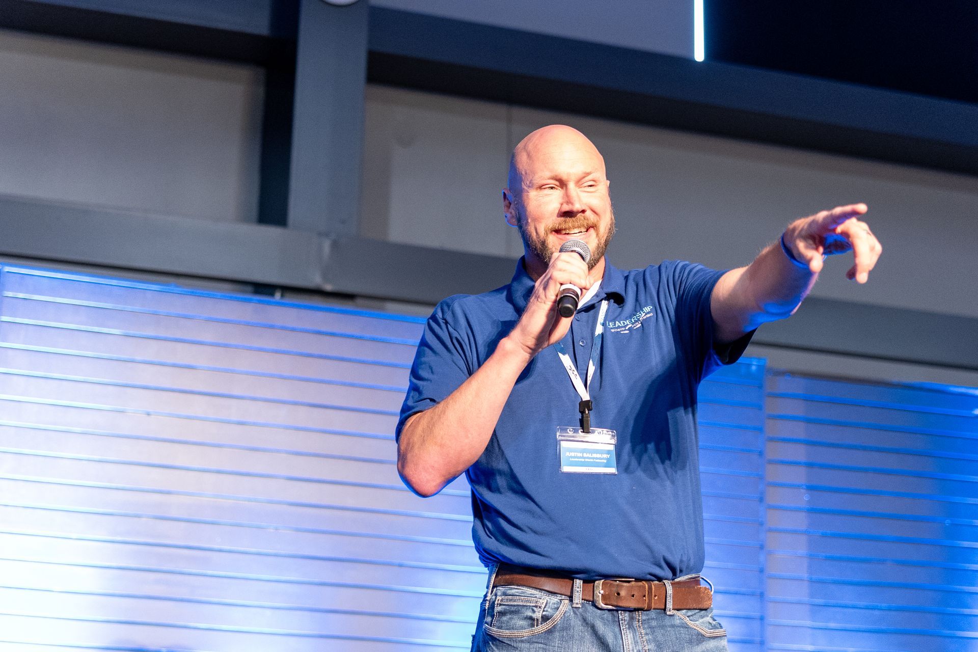 Man with a bald head giving a presentation, pointing to the right, wearing blue shirt and jeans.