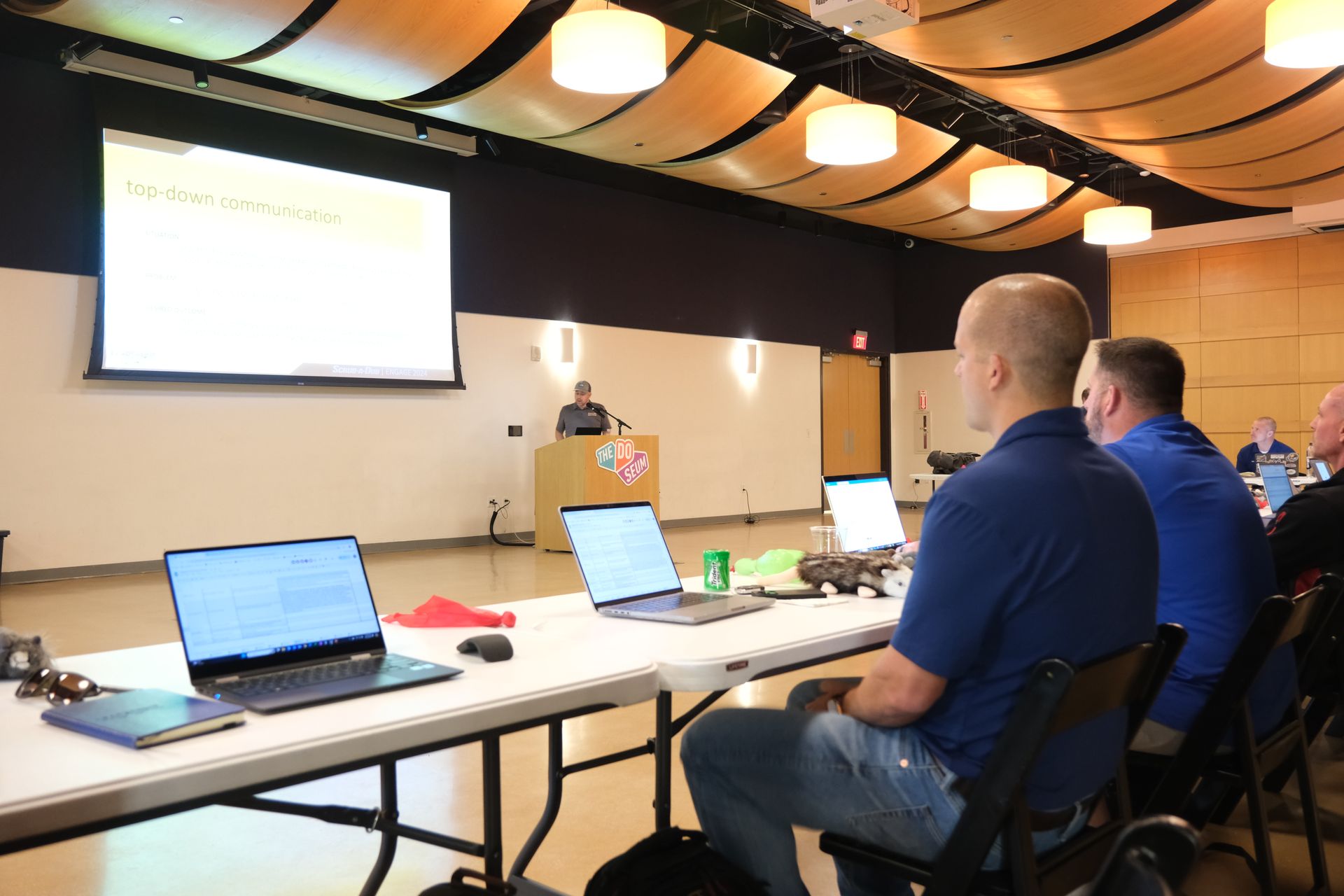 A group of men are sitting at a table with laptops in front of a large screen.