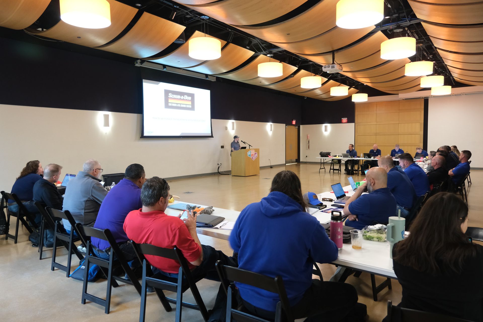 A group of people are sitting at tables in front of a projector screen
