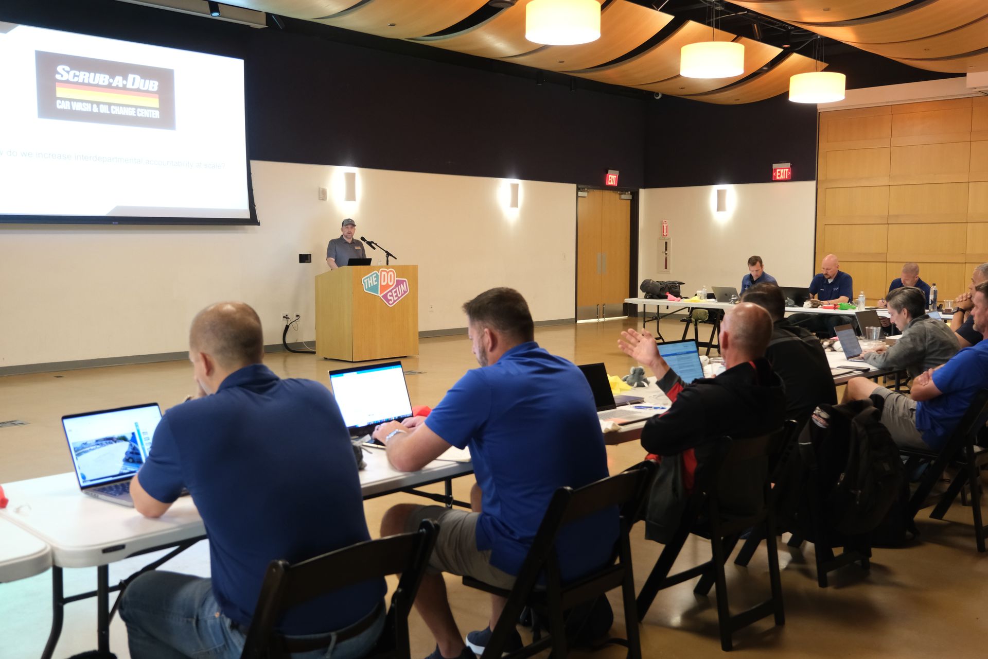 A group of men are sitting at tables in front of a projector screen.