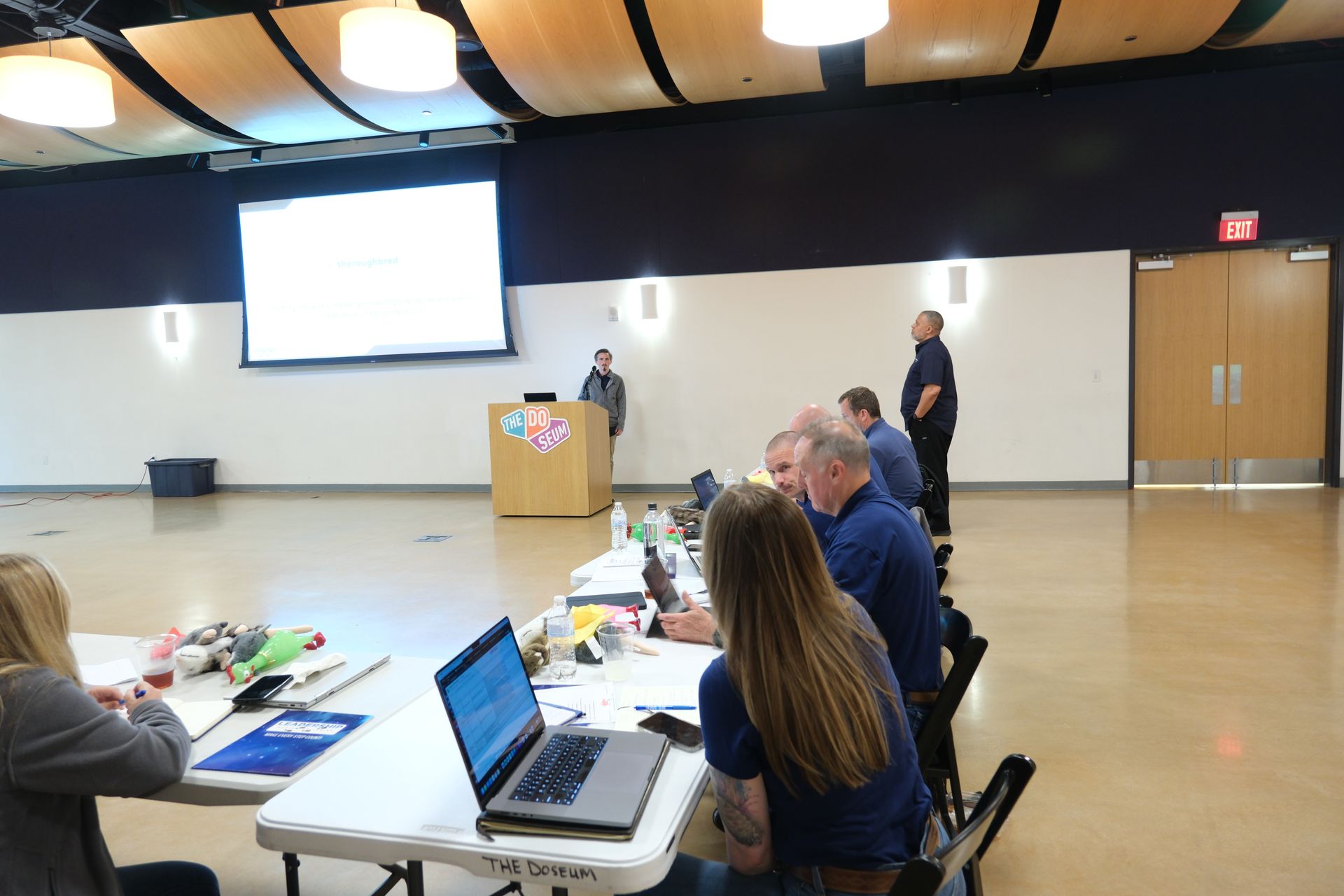 A group of people are sitting at tables with laptops in a large room.
