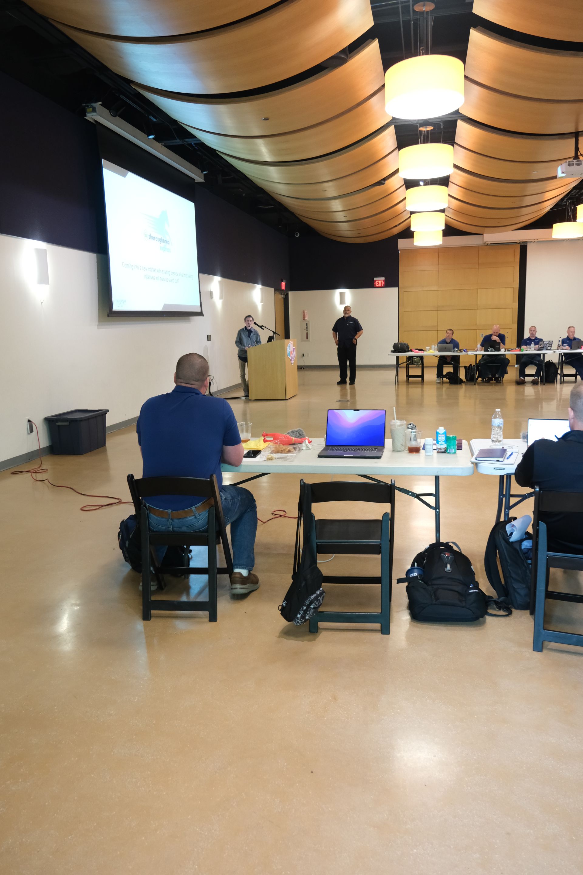 A group of people are sitting at tables in a room with a projector screen.
