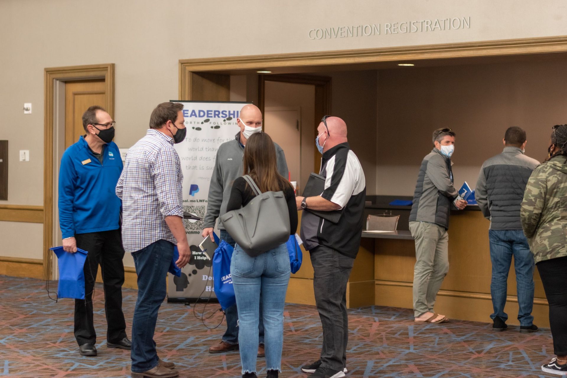 A group of people wearing face masks are standing in a room while checking into the 2020 LWF Conference