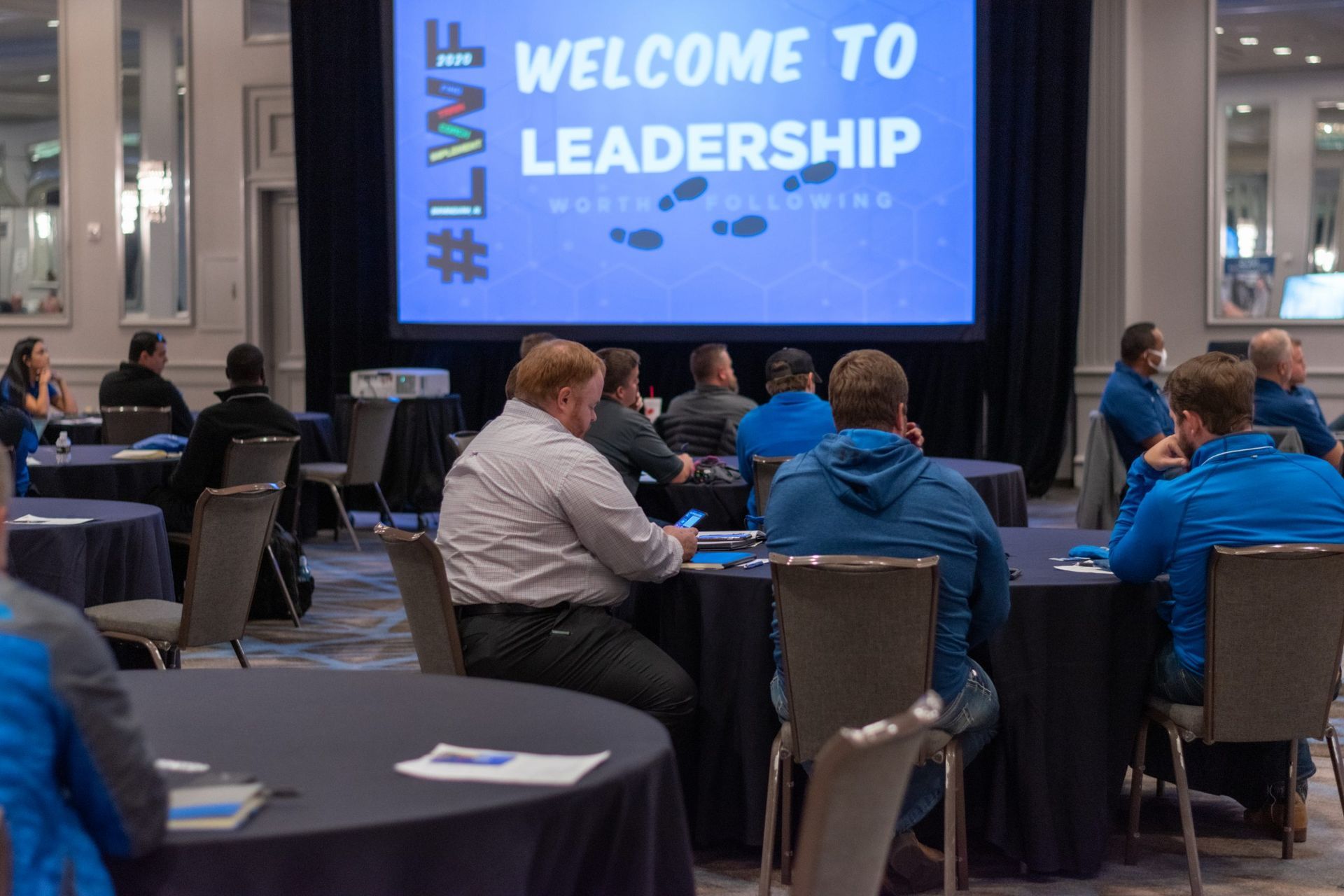 A group of people are sitting at tables in front of a large screen that says welcome to leadership.
