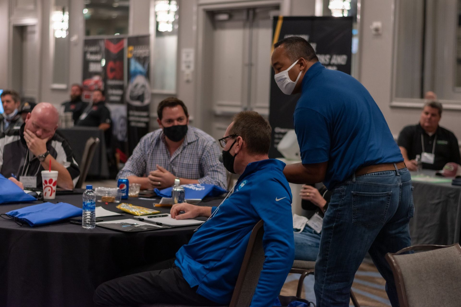 A man wearing a mask is standing next to a man sitting at a table.
