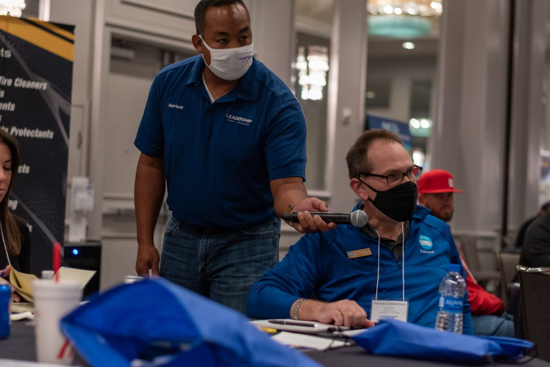 A man wearing a mask is giving a presentation to another man sitting at a table.