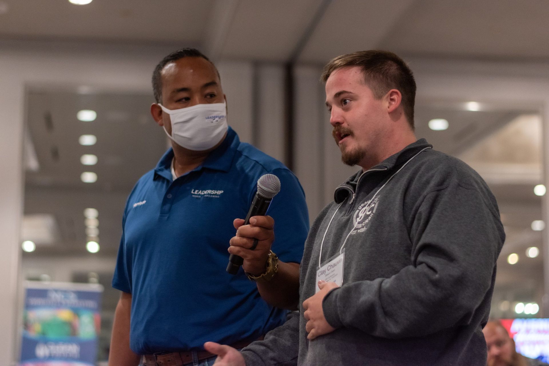 A man wearing a mask is holding a microphone and talking to another man.