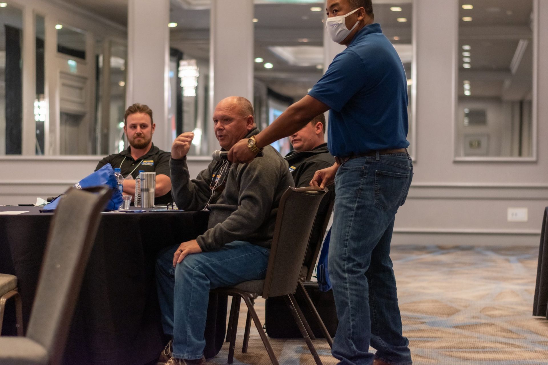 A man wearing a mask is standing next to a group of men sitting at tables.