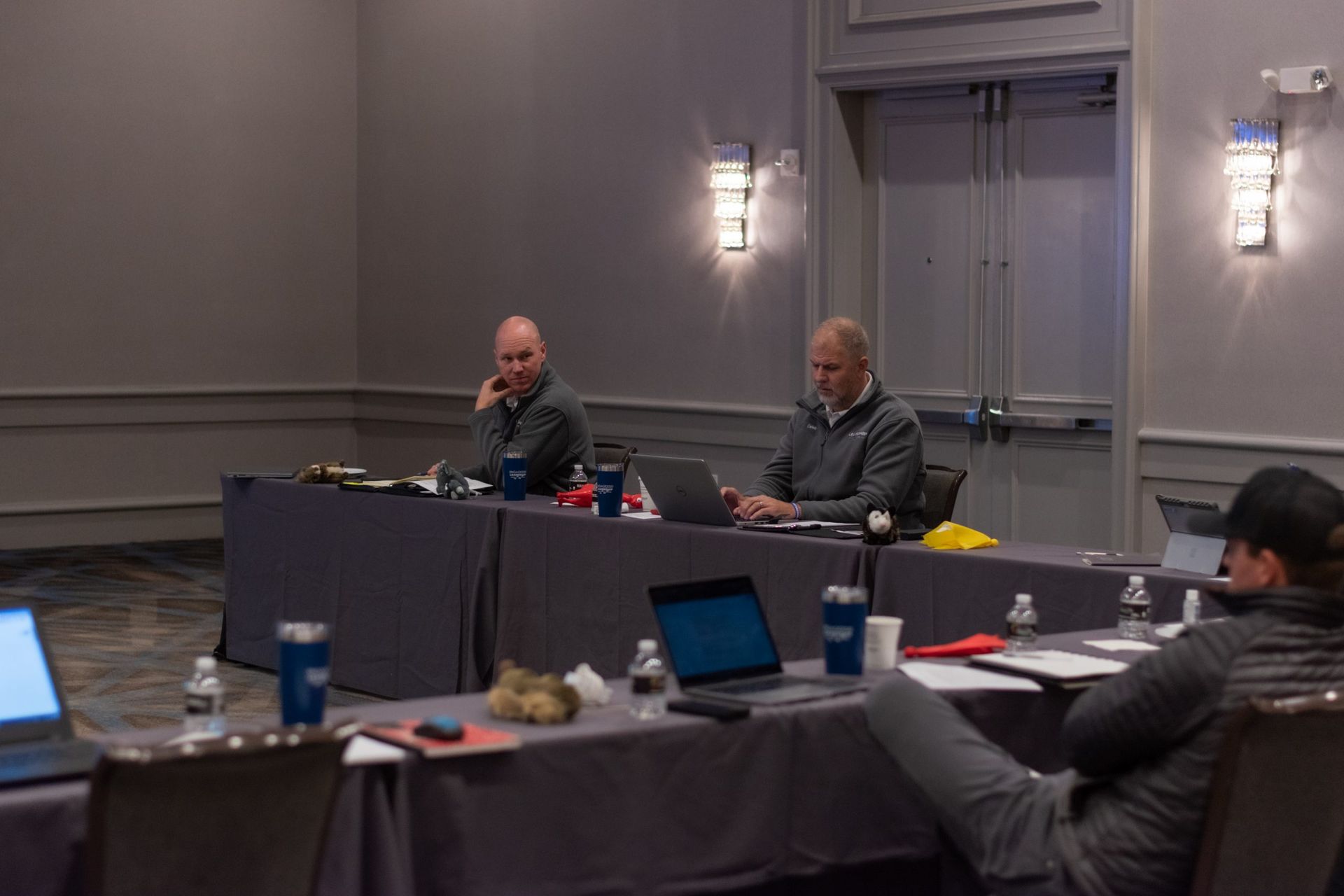 A group of people are sitting at long tables in a conference room with laptops.