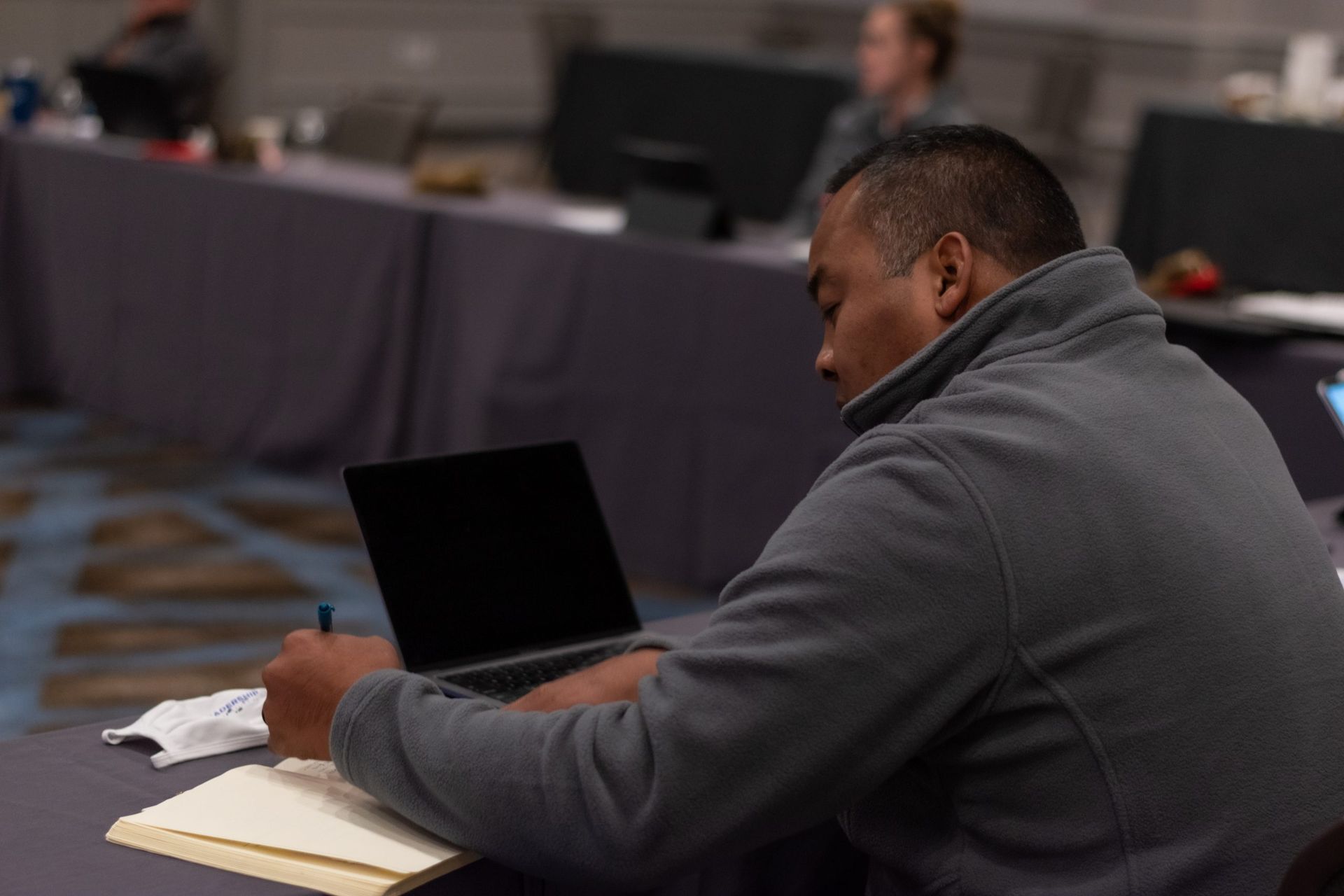 A man is sitting at a table using a laptop and writing in a notebook.