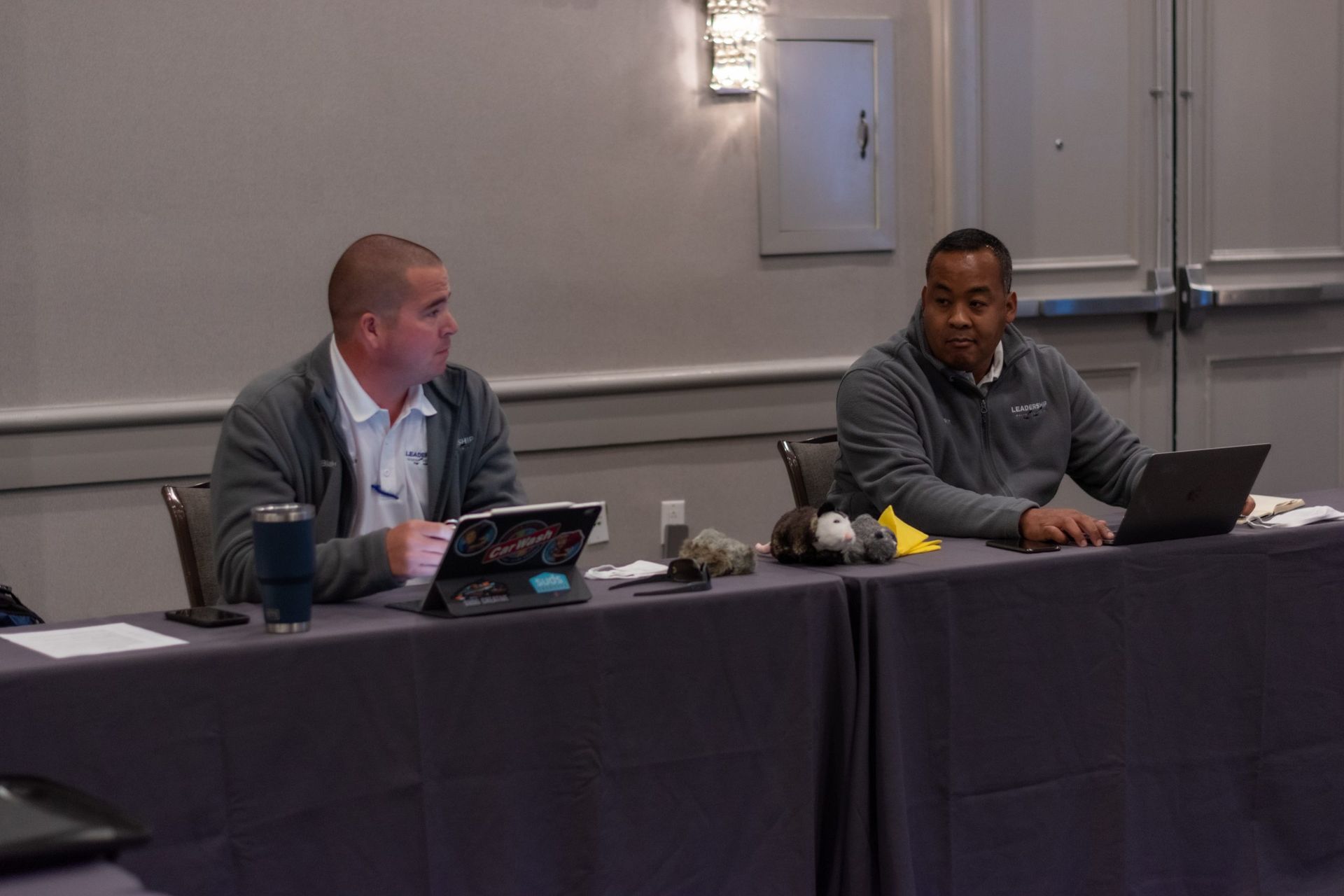Two men are sitting at a long table with laptops.