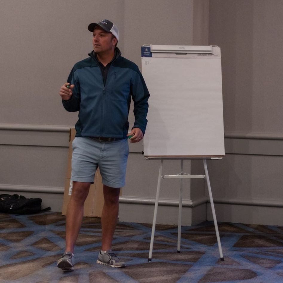 A man in a blue jacket stands in front of a white board