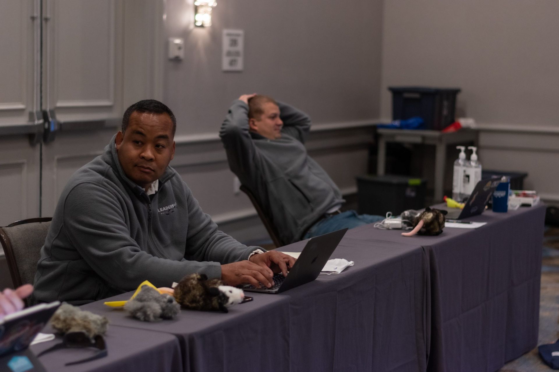 Two men are sitting at a long table using laptops.
