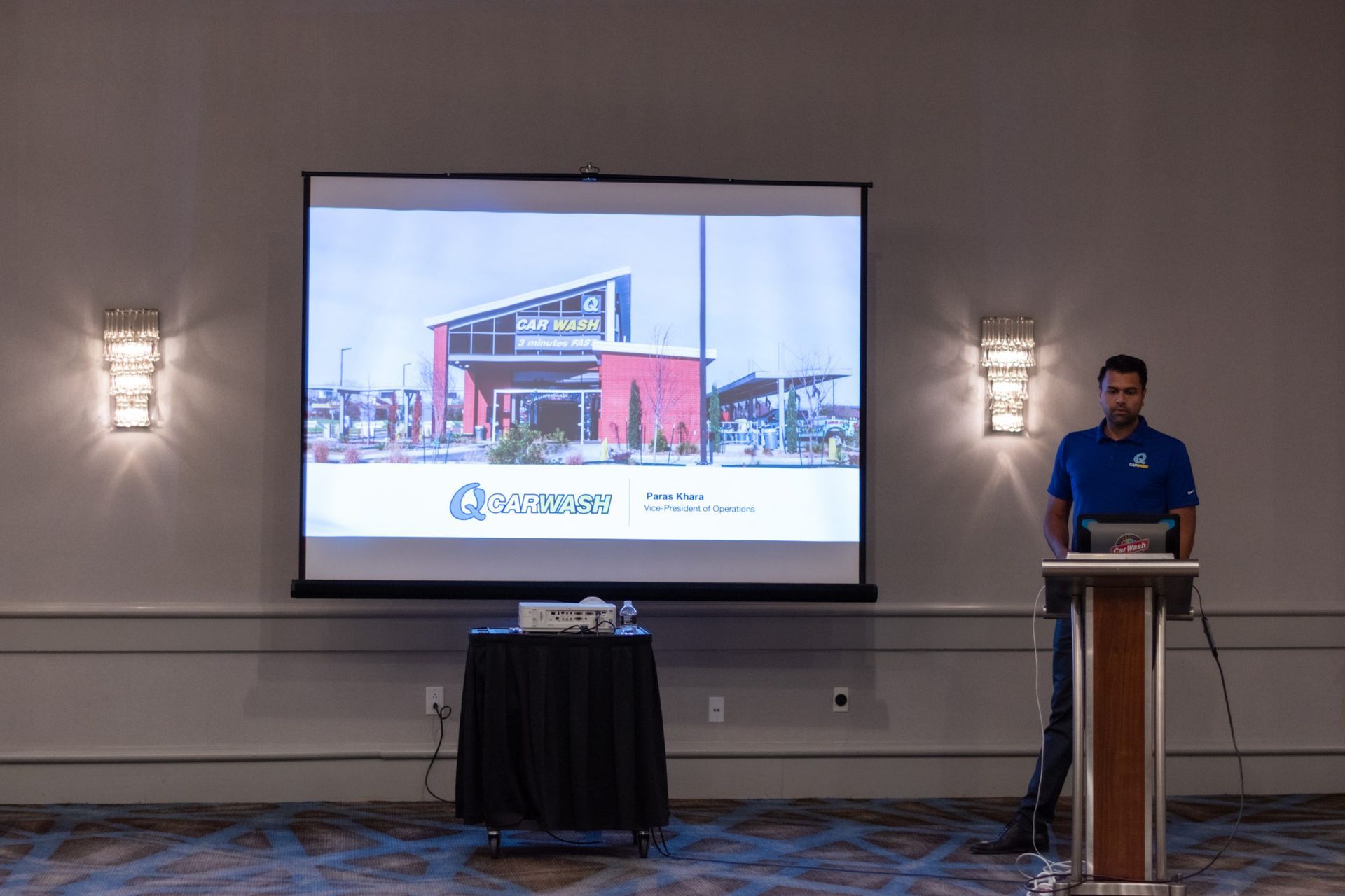 A man is standing at a podium in front of a large screen.