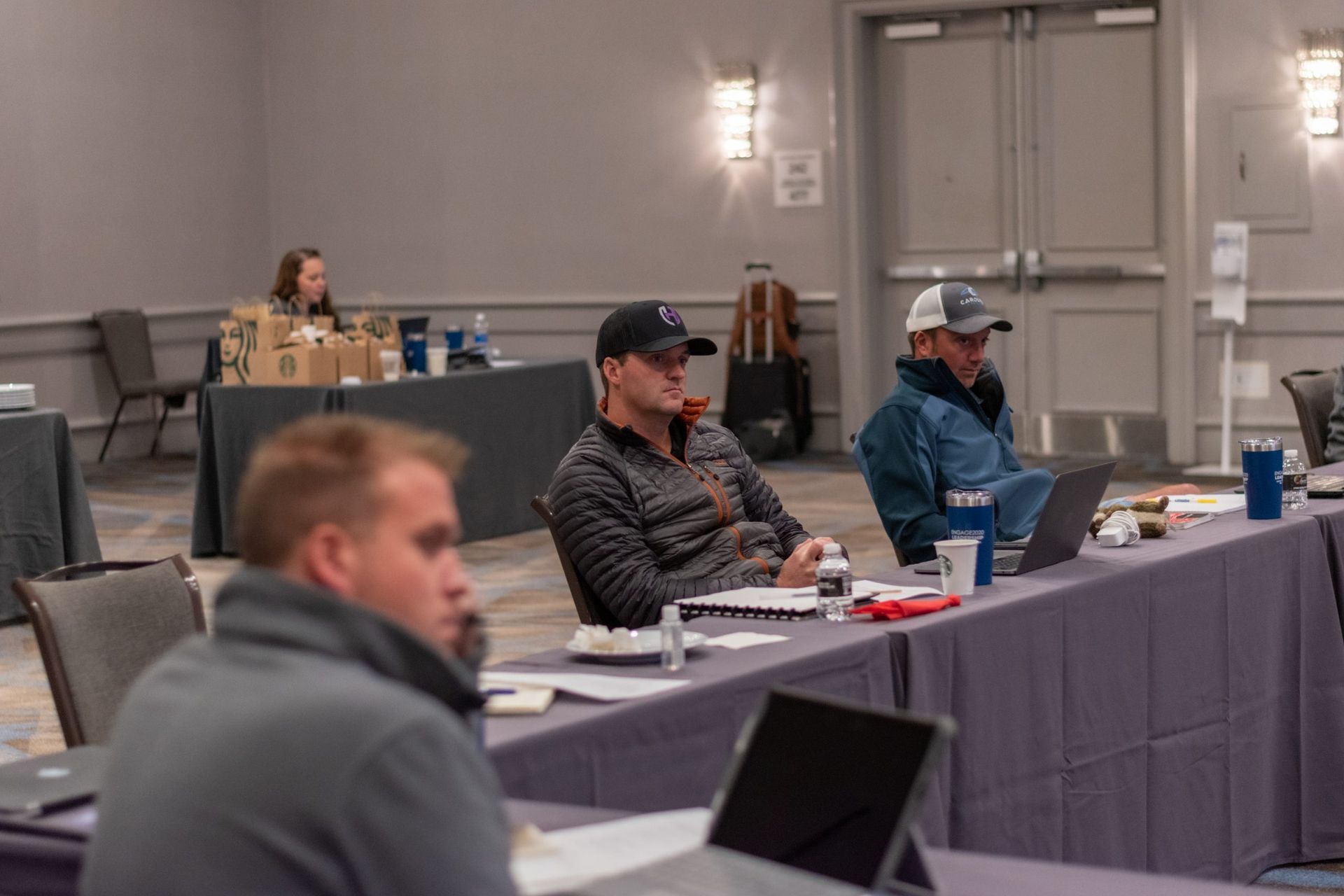A group of men are sitting at long tables with laptops in a conference room.
