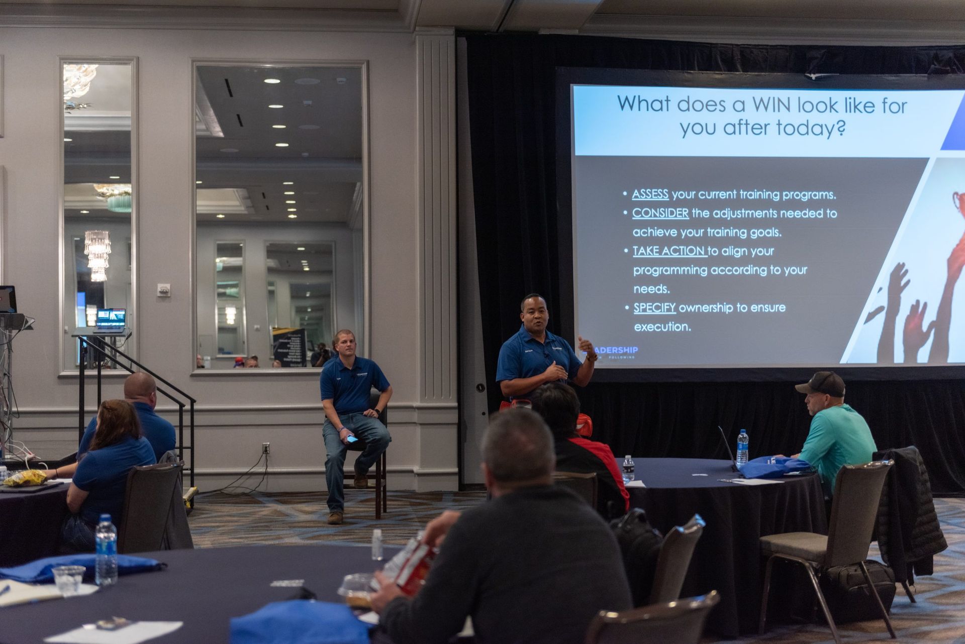 A group of people are sitting at tables in front of a large screen.