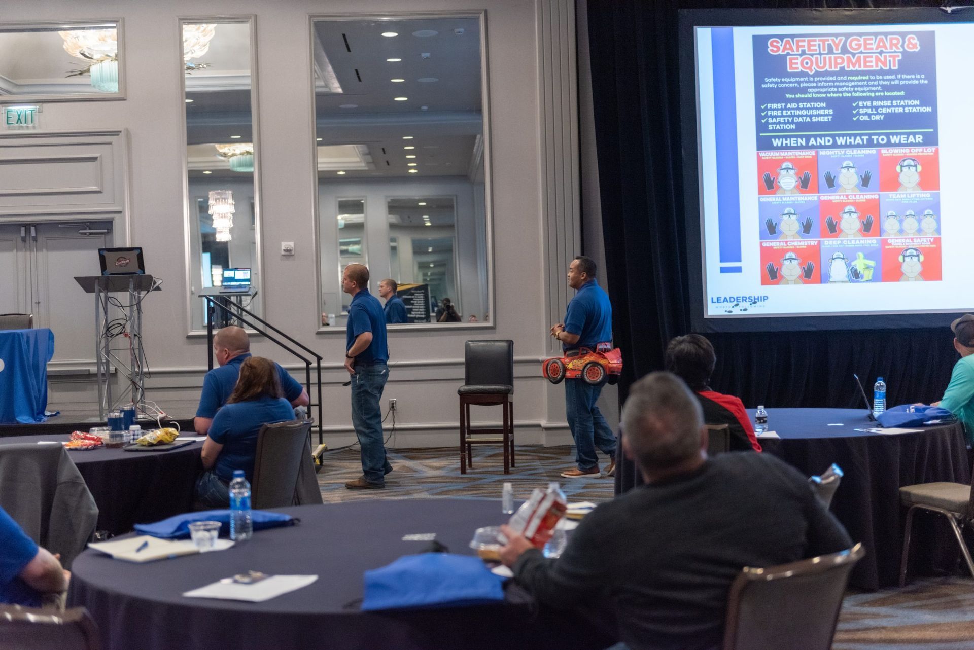 A group of people are sitting at tables in front of a projector screen.